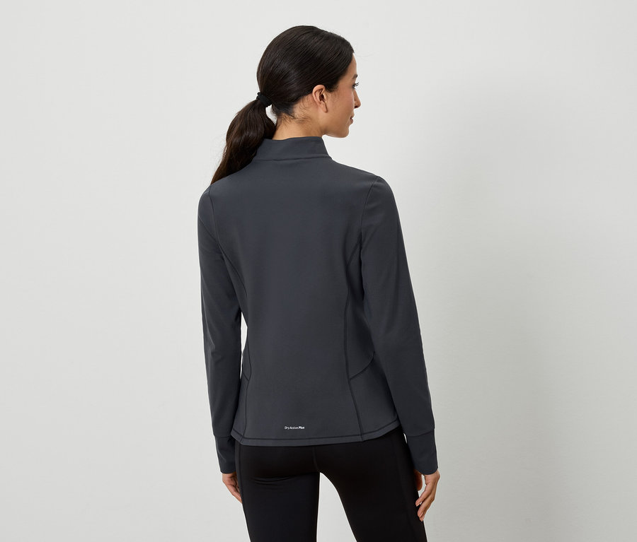 Kobieta z brązowymi włosami związanymi w kucyk pozuje tyłem do aparatu. Ma na sobie szarą kurtkę sportową i czarne legginsy.