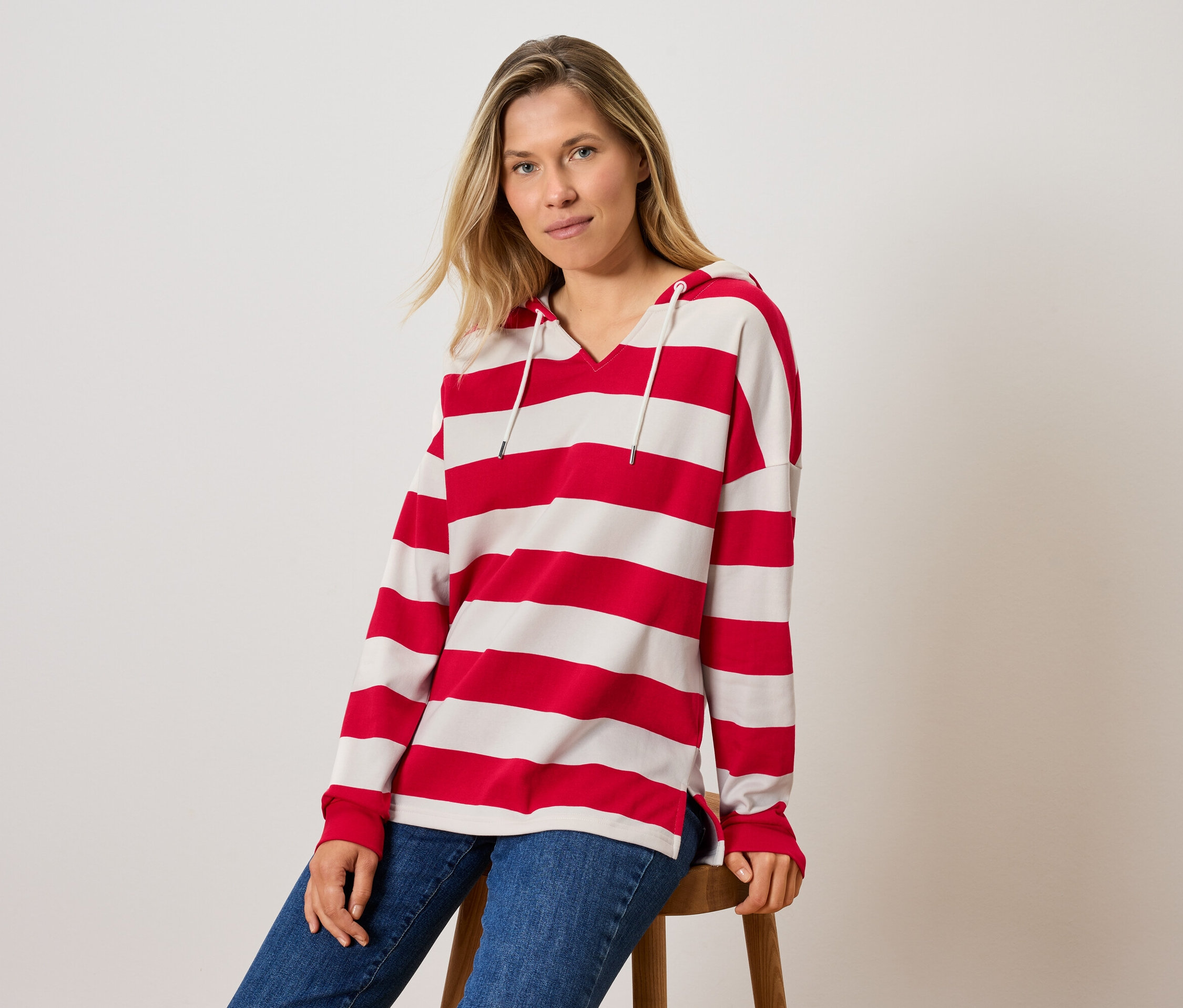 Kobieta siedzi na stołku, ubrana w bluzę w paski z kapturem i dżinsy.