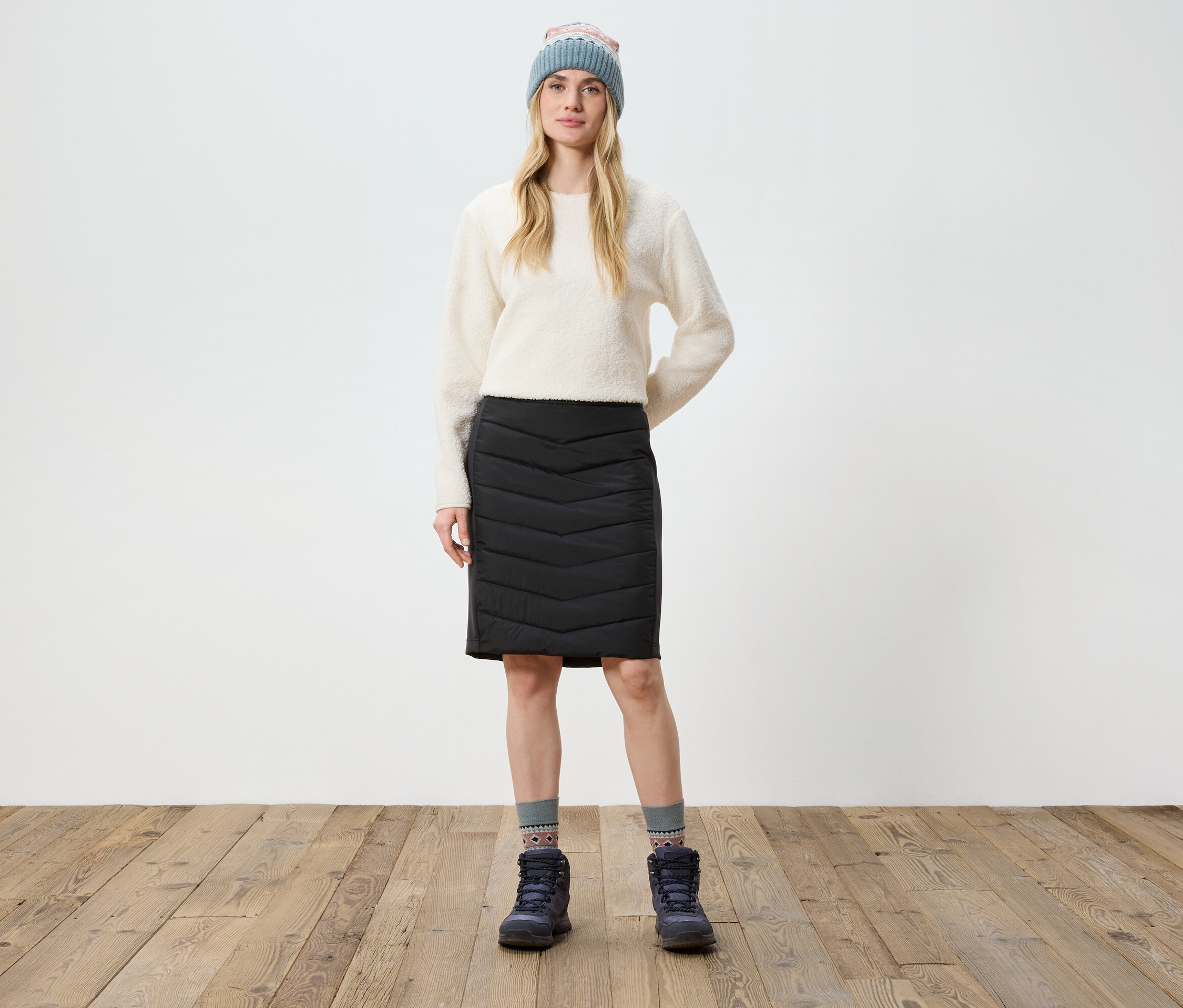 Kobieta stoi na drewnianej podłodze, ubrana w kremowy sweter, czarną spódnicę, buty i dzianinową czapkę.