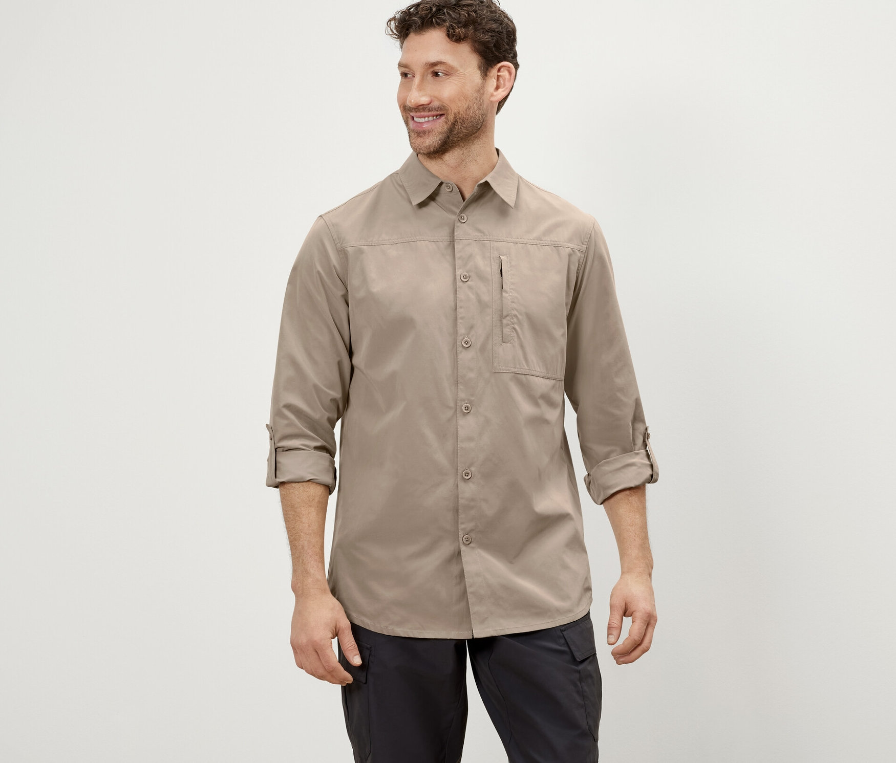 Mężczyzna pozuje w beżowej koszuli outdoorowej z ochroną przed komarami i czarnych spodniach.