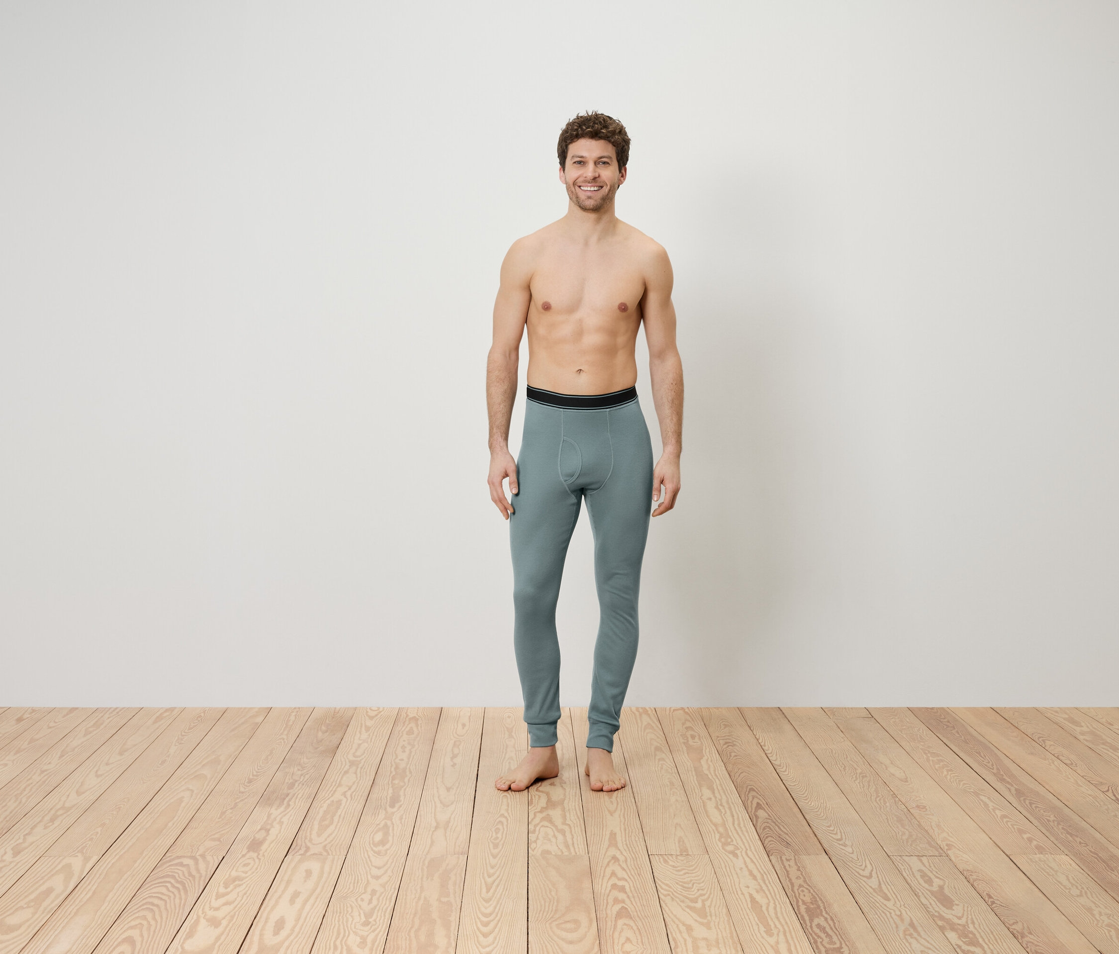 Mężczyzna z odkrytą górną częścią ciała stoi na drewnianej podłodze, ubrany w szare legginsy.
