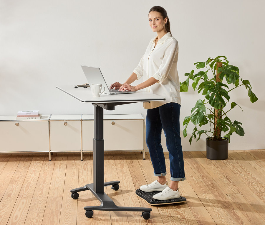 Kobieta stoi przy biurku z regulowaną wysokością i korzysta z laptopa, obok stoi kubek.