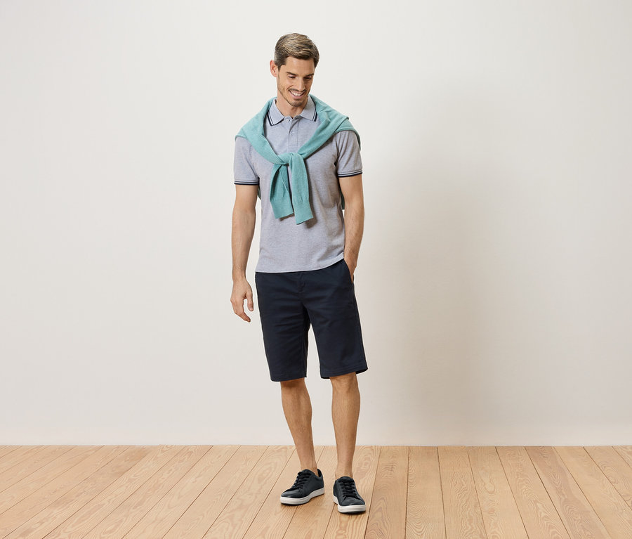 Mężczyzna stoi z jasnozielonym swetrem na szyi, szarą koszulką polo, granatowymi szortami i niebieskimi tenisówkami.