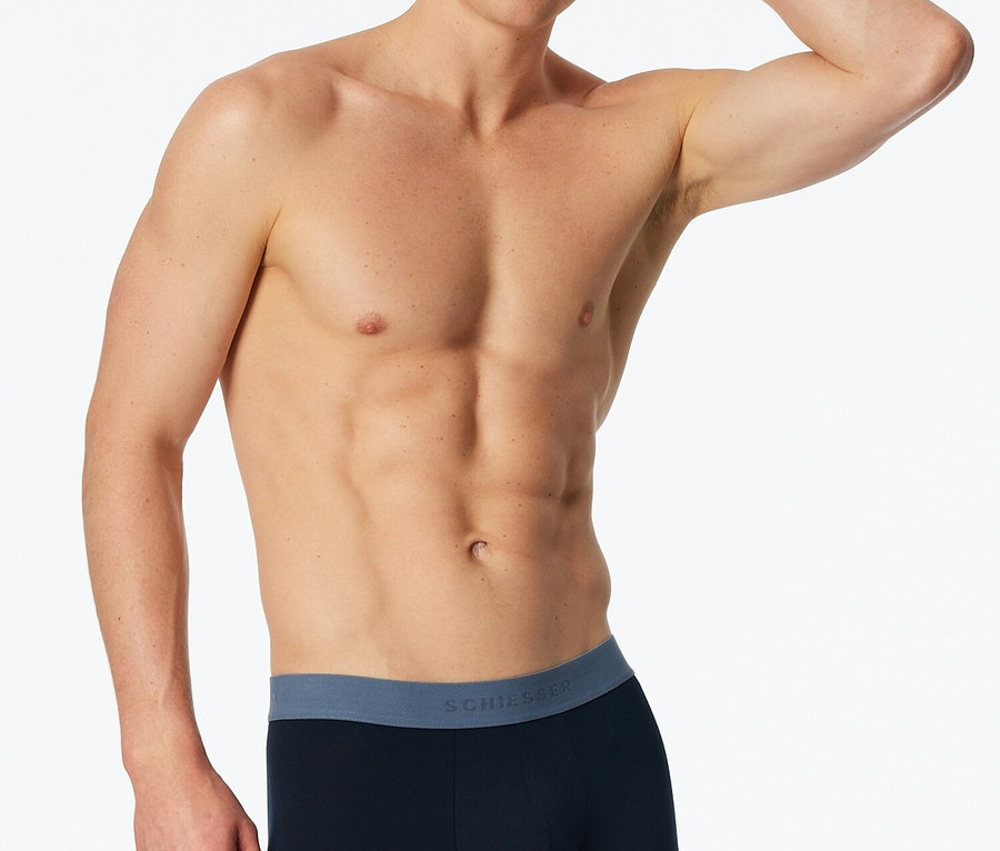 Mężczyzna bez koszuli pozuje w granatowych bokserkach z niebieskim pasem na białym tle.