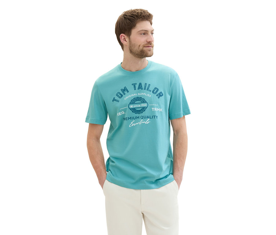 Mężczyzna w koszulce z logo Tom Tailor, meadow teal i beżowych spodniach z rękami w kieszeniach.