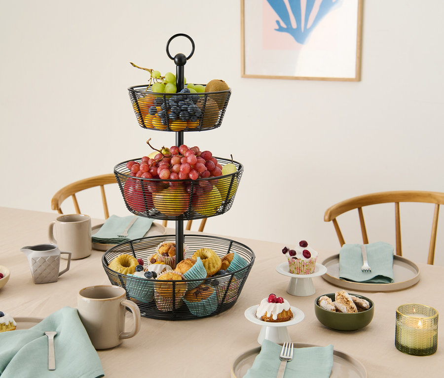 Na stole stoi metalowy, trzypoziomowy kosz na owoce z ciastami, owocami i naczyniami.