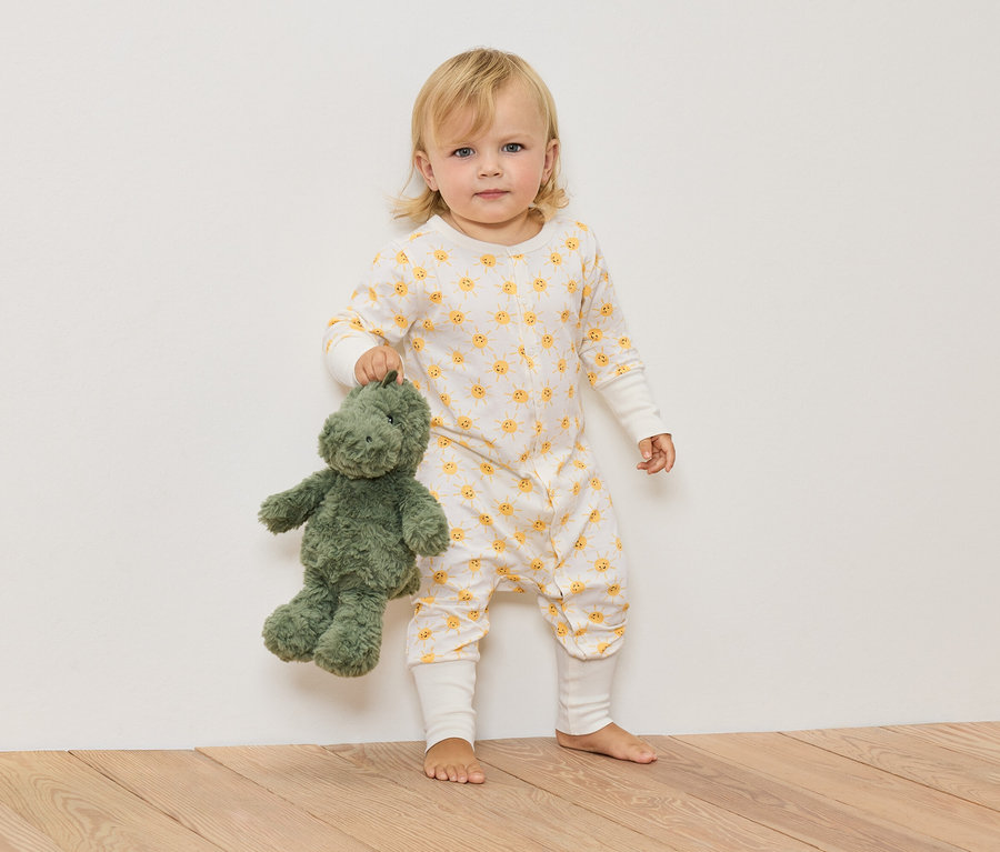 Blondwłose dziecko w kombinezonie w słoneczka trzyma zielonego pluszowego dinozaura.