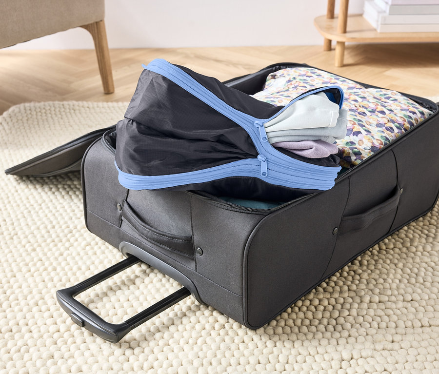 Otwarty, szary kufer na dywanie, wypełniony ubraniami i czarną kostką do pakowania z niebieskim zamkiem błyskawicznym.