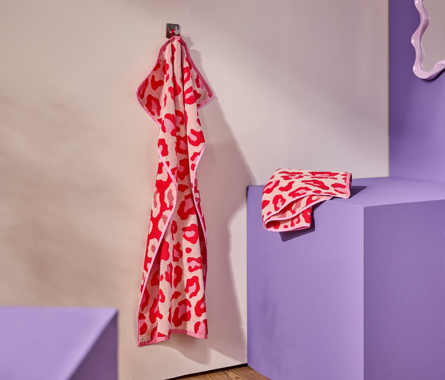 Czerwono-różowy ręcznik w panterkę wisi na haku. Złożony ręcznik leży na fioletowym postumencie.