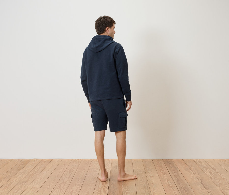 Mężczyzna od tyłu, ubrany w granatową bluzę z kapturem i szorty na drewnianej podłodze.