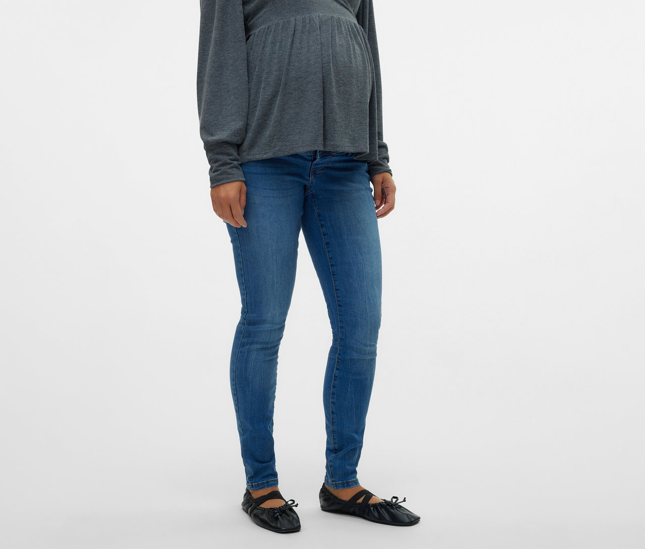 Kobieta w ciąży ma na sobie szary top i dżinsy skinny MAMALICIOUS „MLMILA” oraz czarne baleriny.