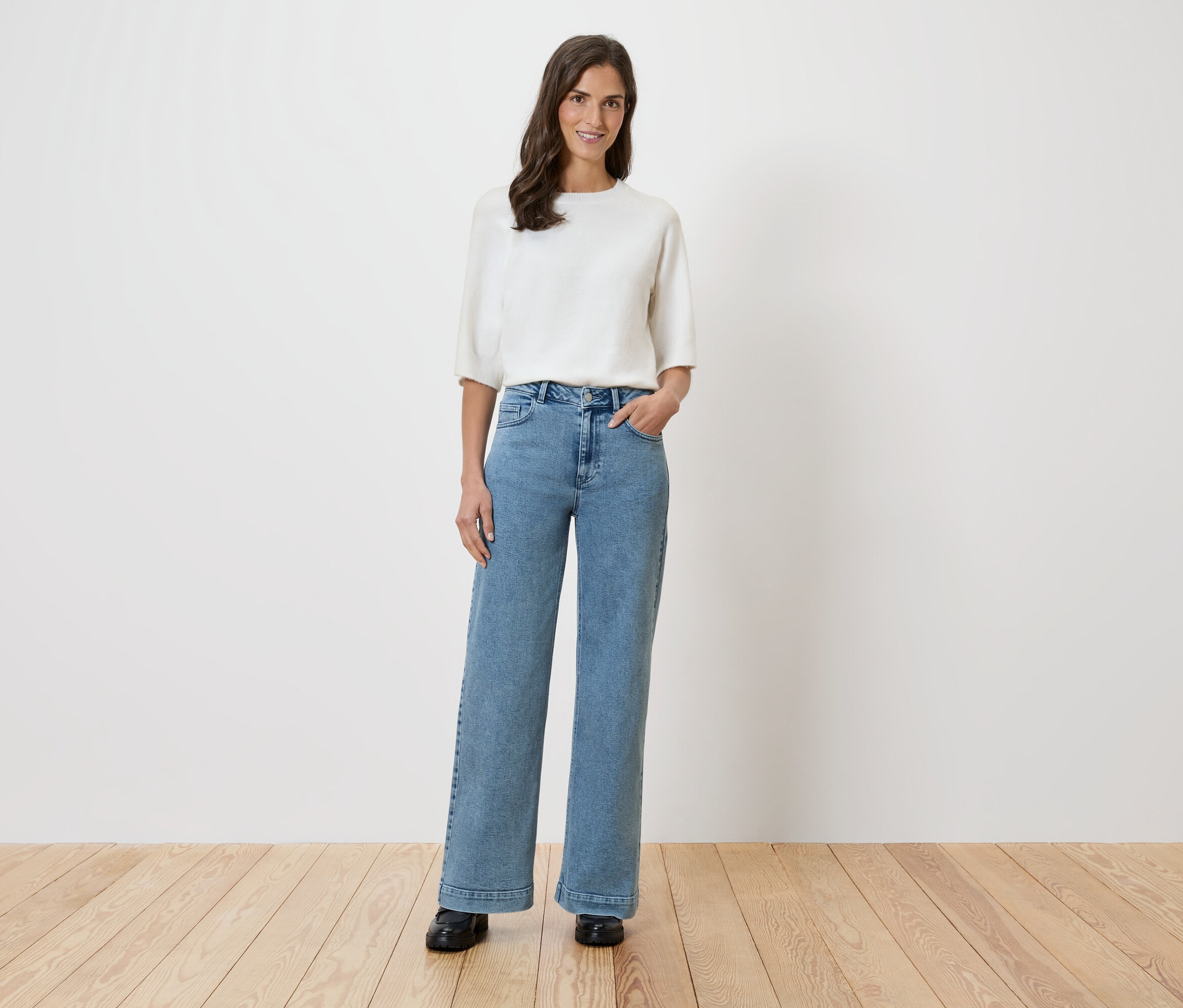 Kobieta stoi na tle białej ściany, ubrana w białą bluzkę i szerokie dżinsy.