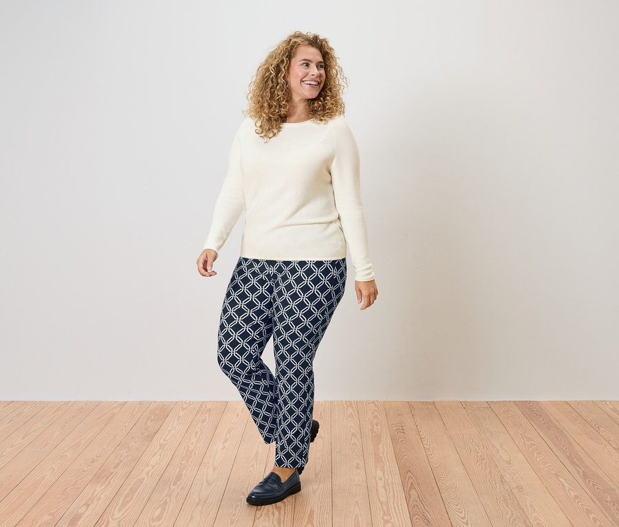 Kobieta z kręconymi włosami w kremowym swetrze i granatowych spodniach w geometryczny wzór.