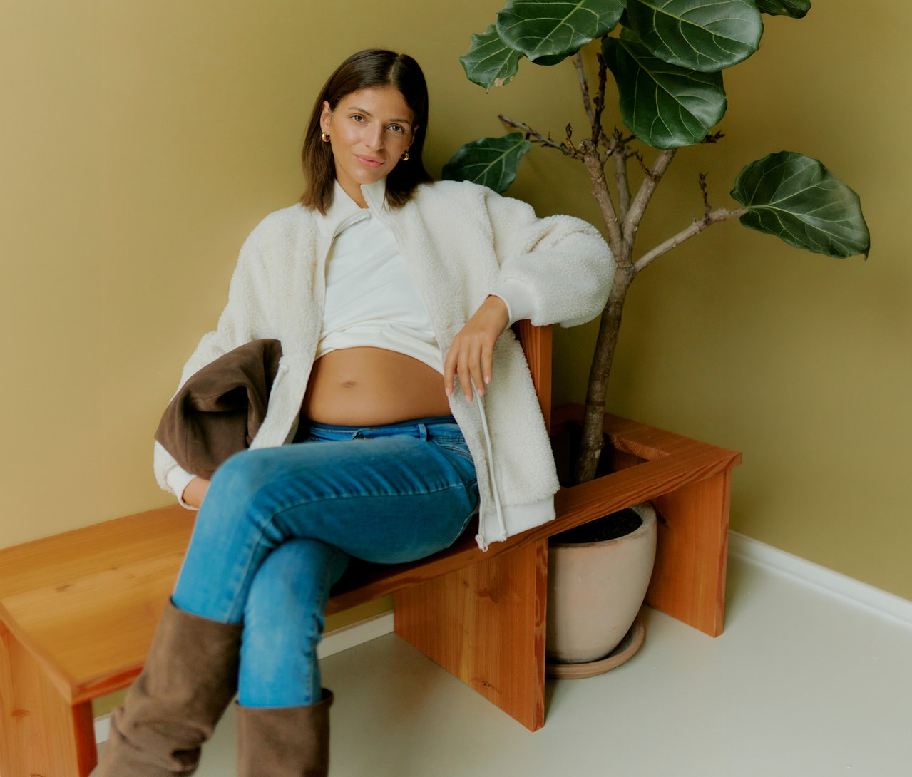 Kobieta w ciąży siedzi na ławce obok rośliny, ma na sobie rozpiętą pluszową bluzę 3 w 1 MAMALICIOUS „MLCARLY”, białą bluzkę i dżinsy.