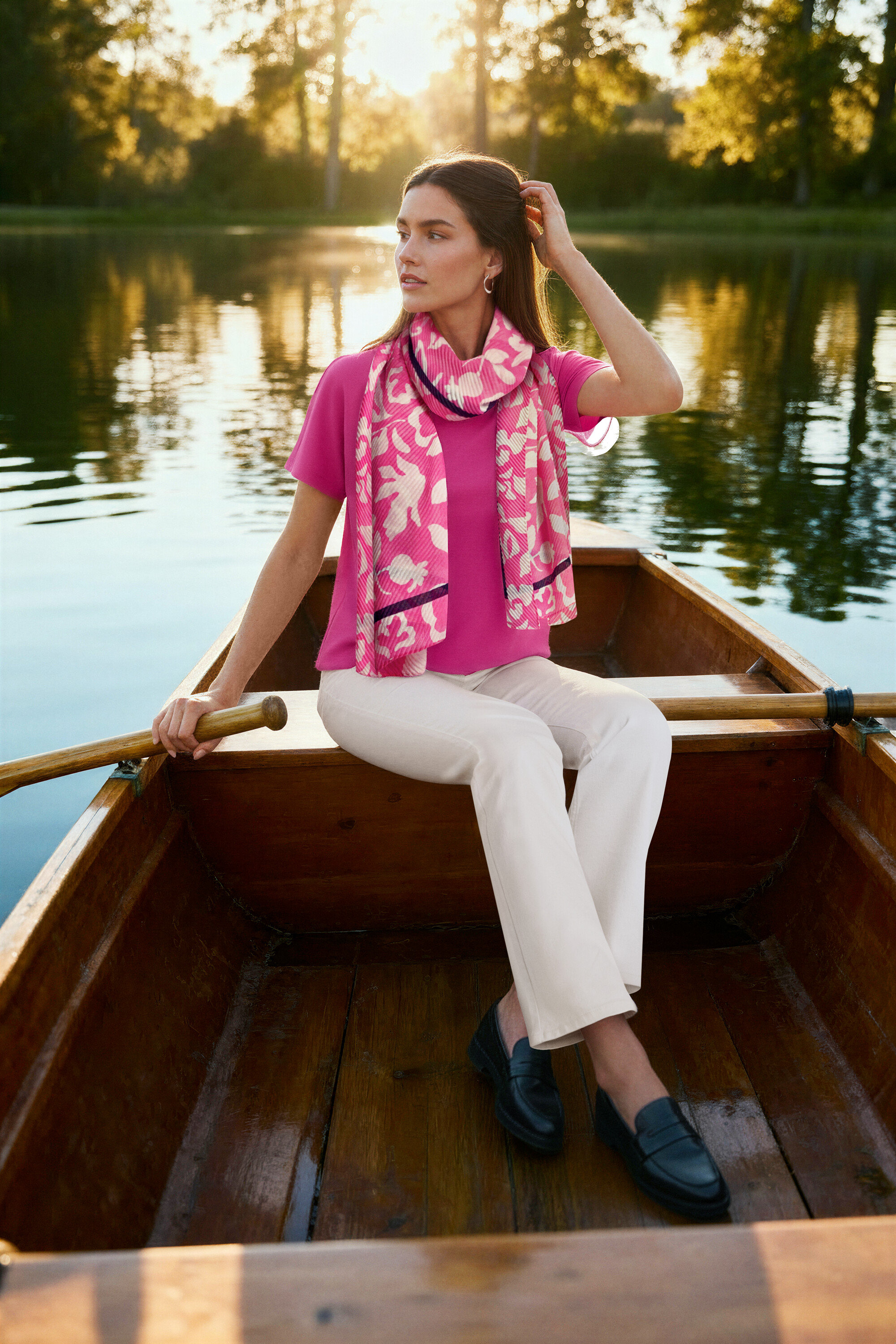 Kobieta siedzi w łodzi, ma na sobie różową bluzkę, kremowe bootcut jeansy ‒ Fit »Lou«, mokasyny skórzane i kolczyki kreole Wave.