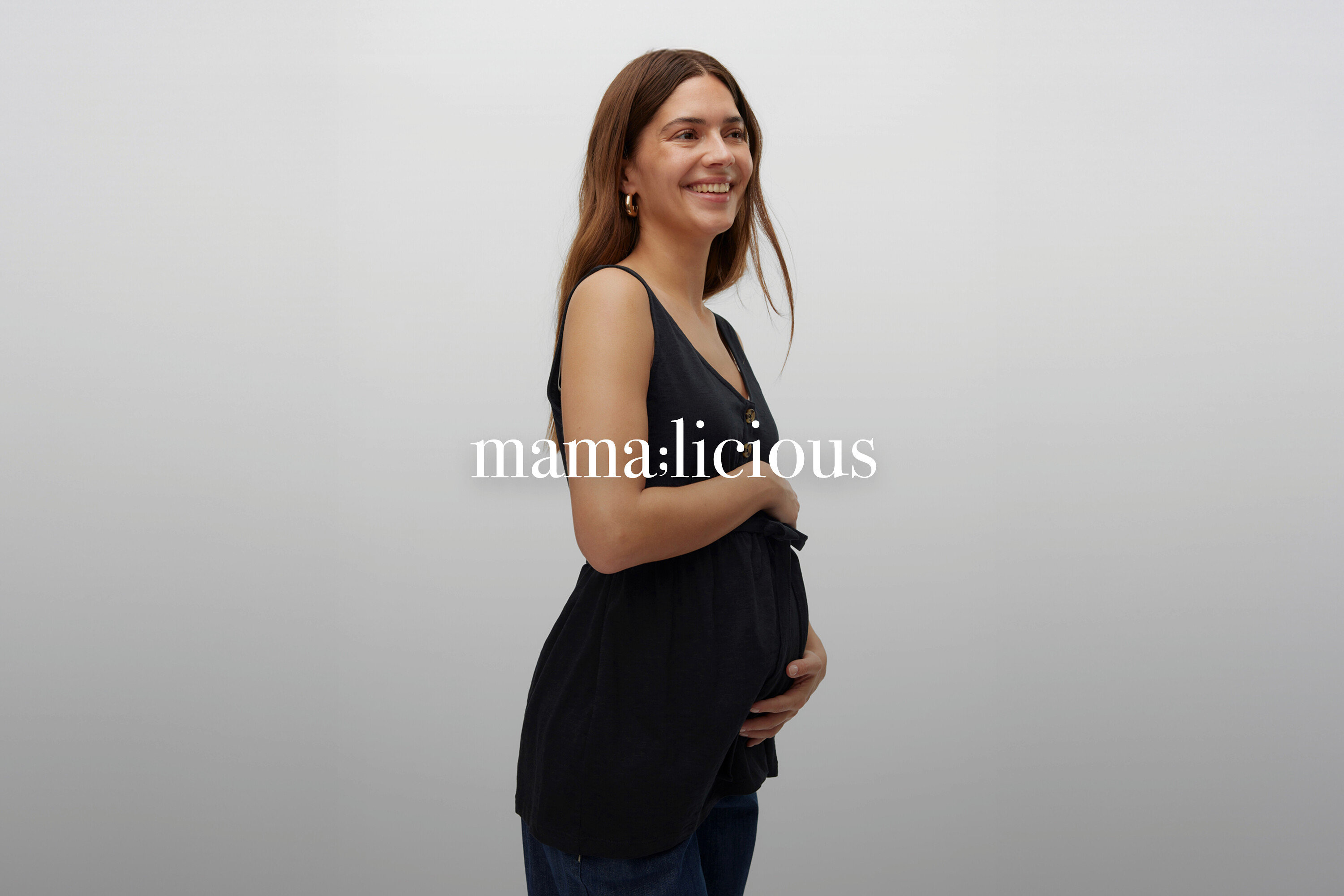 Uśmiechnięta kobieta w ciąży z długimi brązowymi włosami w czarnym topie bez rękawów i dżinsach.