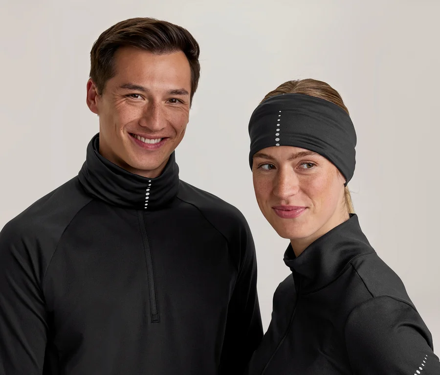 Uśmiechnięci mężczyzna i kobieta w czarnych sportowych bluzkach z wysokimi kołnierzami i odblaskowymi kropkami. Kobieta ma też czarną opaskę na głowę z odblaskowymi kropkami.