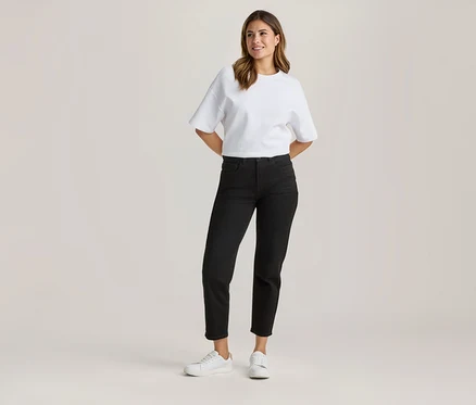Stojąca kobieta w białym, luźnym T-shircie, czarnych dżinsach 7/8 i białych sneakersach, z rękami za plecami, uśmiechnięta.