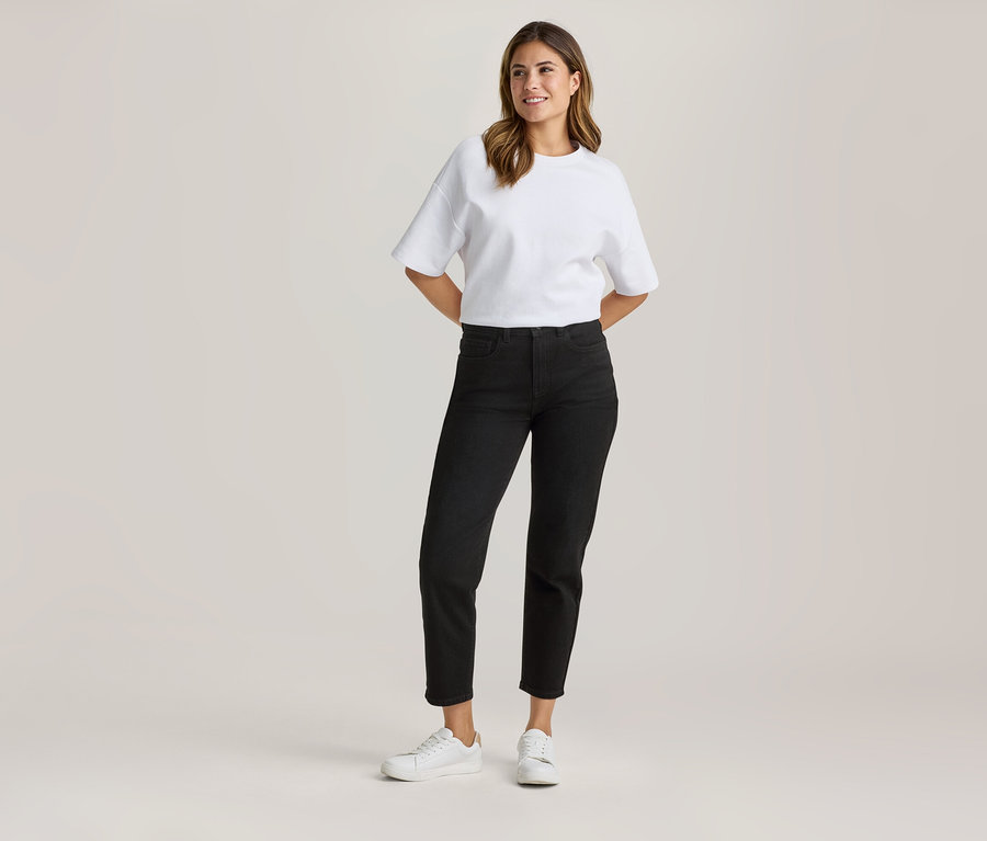 Stojąca kobieta w białym, luźnym T-shircie, czarnych dżinsach 7/8 i białych sneakersach, z rękami za plecami, uśmiechnięta.