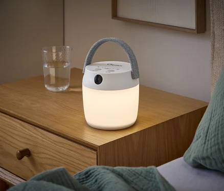 Biała lampa LED z szarym uchwytem świeci na drewnianej szafce nocnej obok szklanki wody.