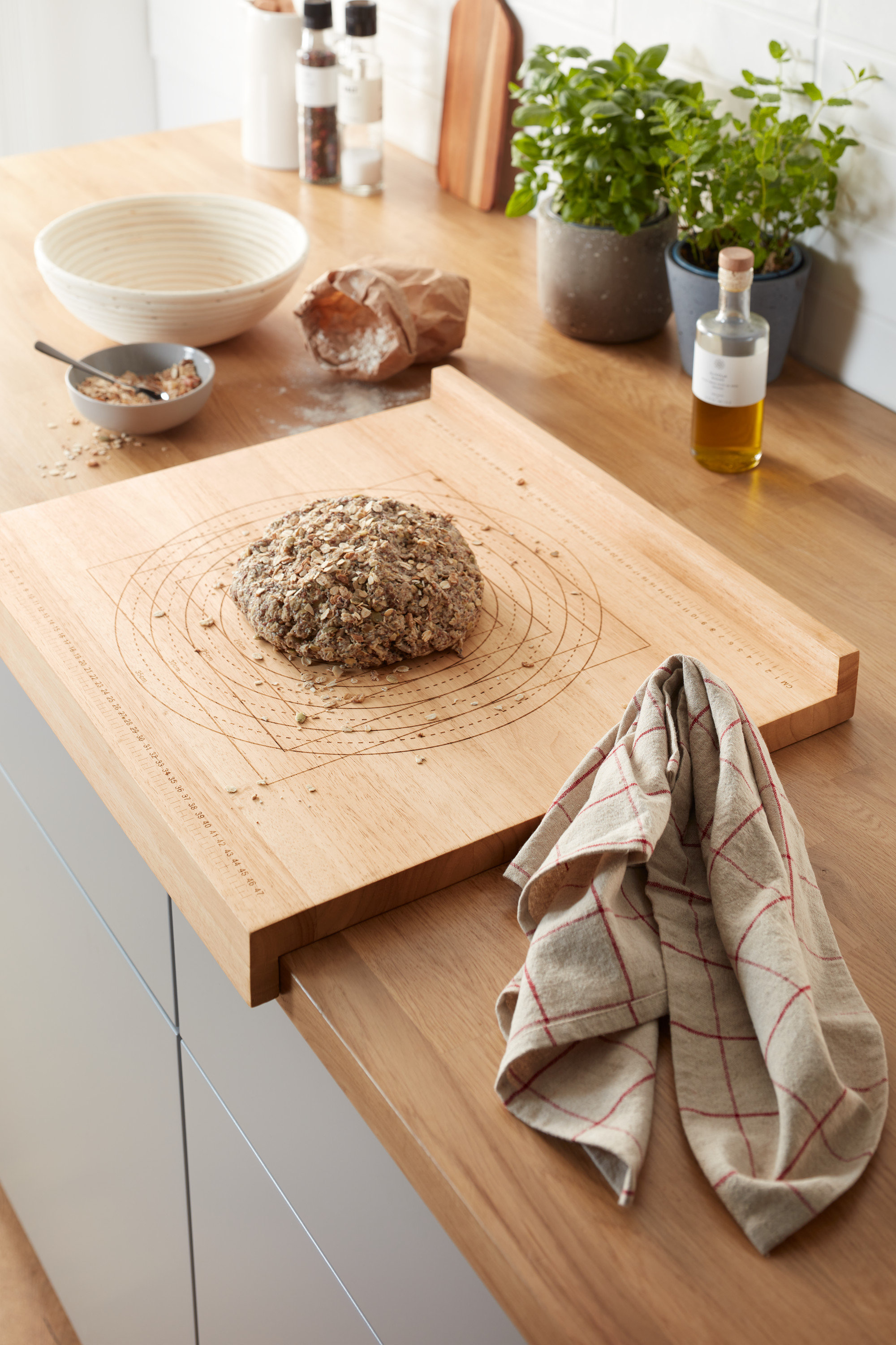 Na blacie kuchennym leży drewniana deska do prac kuchennych i przygotowywania wypieków z bochenkiem chleba. W tle zioła w doniczkach i inne przybory kuchenne.