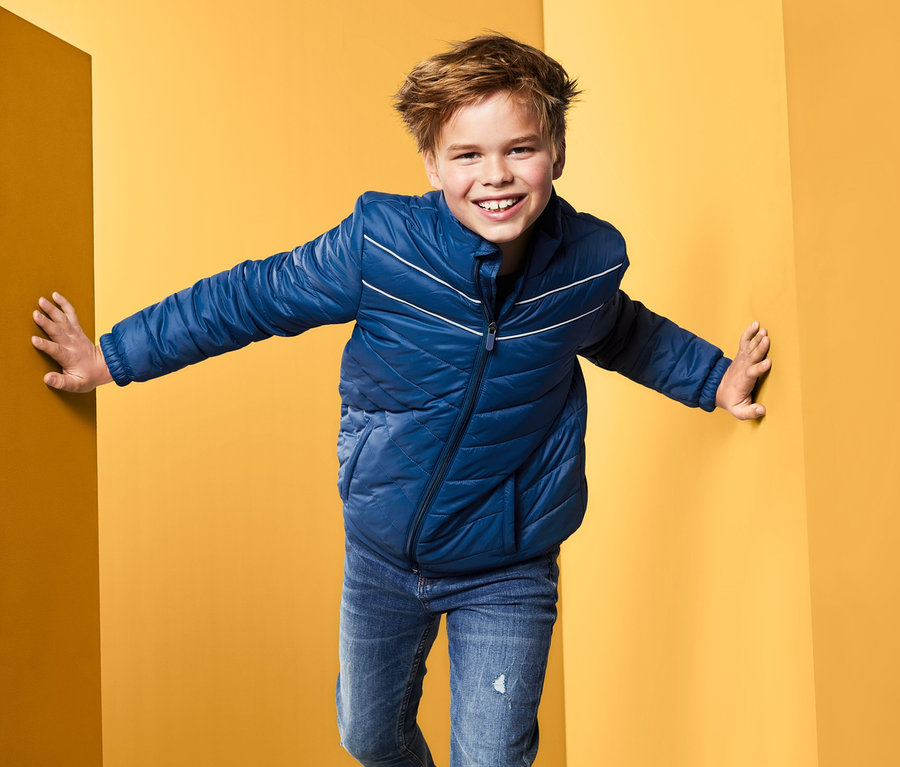 Chłopiec w niebieskiej kurtce pikowanej stoi w rogu żółtego pokoju.