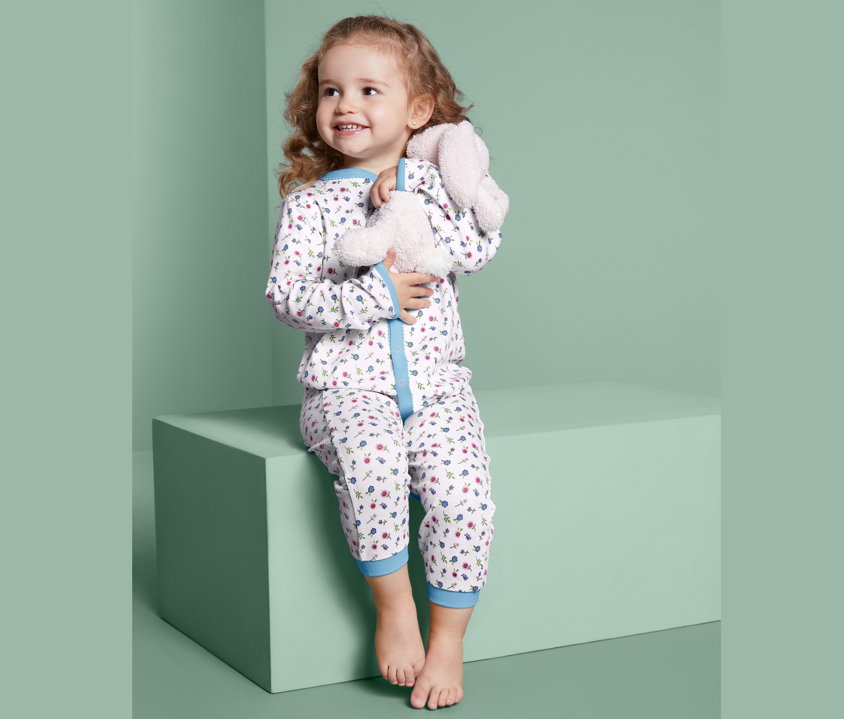 Dziewczynka siedzi na bloku i przytula pluszowego króliczka. Ma na sobie Śpioszki pajacyki bawełniane z długimi rękawami, 2 sztuki.