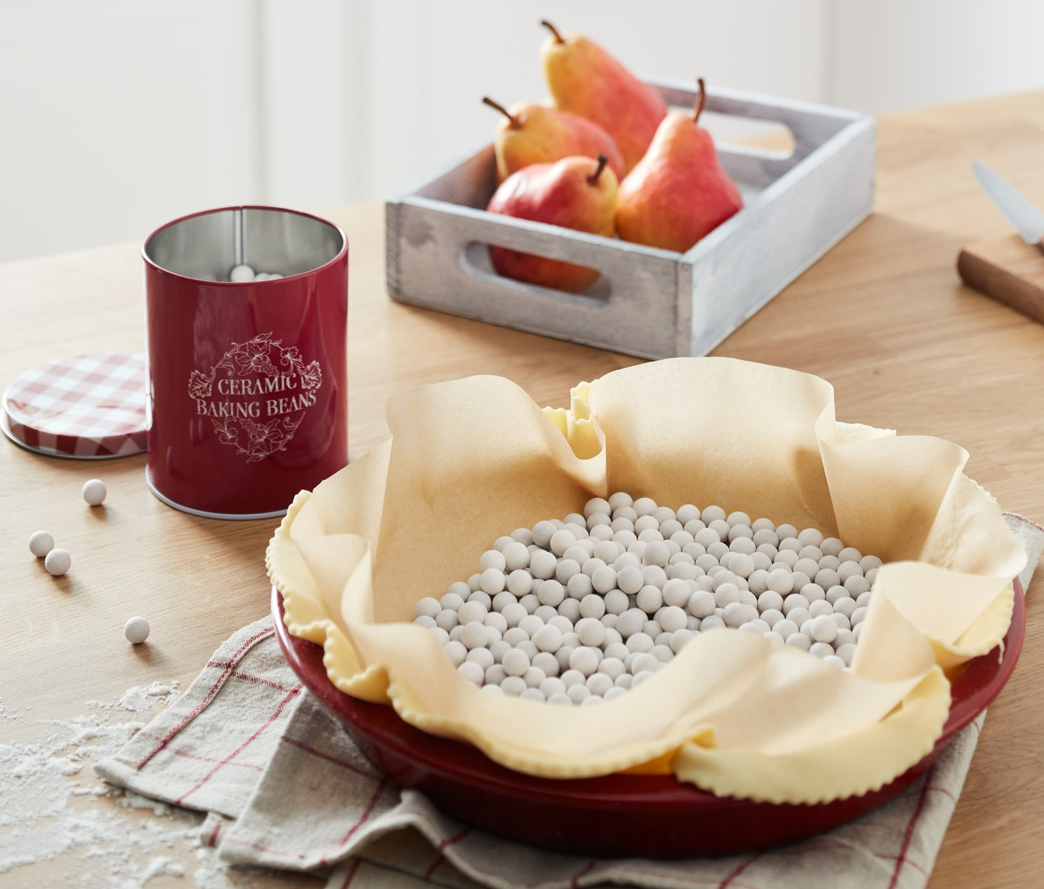 Czerwona forma do ciasta z ciastem i grochem ceramicznym. Puszka grochu ceramicznego i skrzynka gruszek na drewnianym stole.