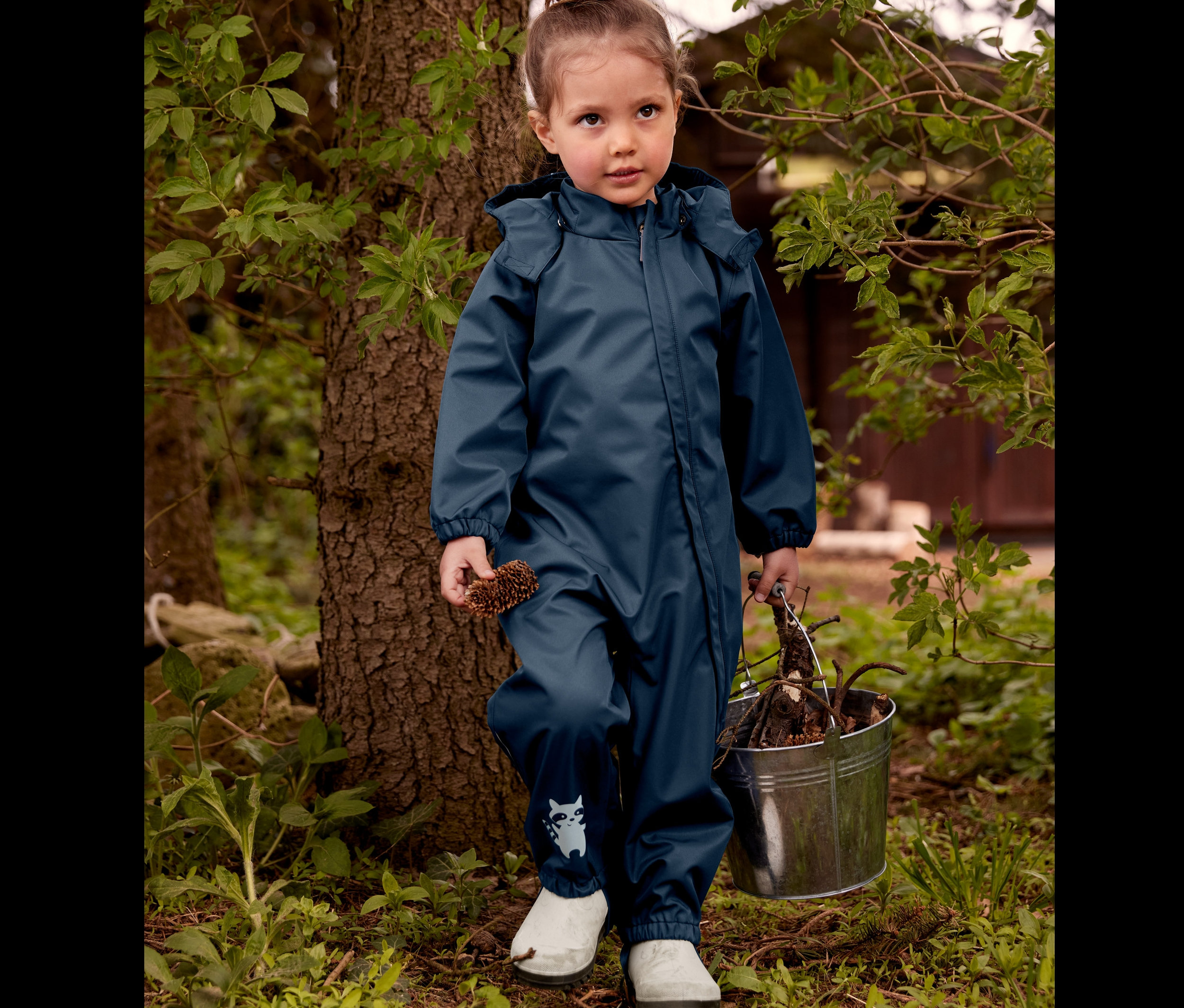 Dziewczynka stoi przy drzewie w kaloszach i kombinezonie przeciwdeszczowym, trzyma szyszkę i wiadro z patykami.