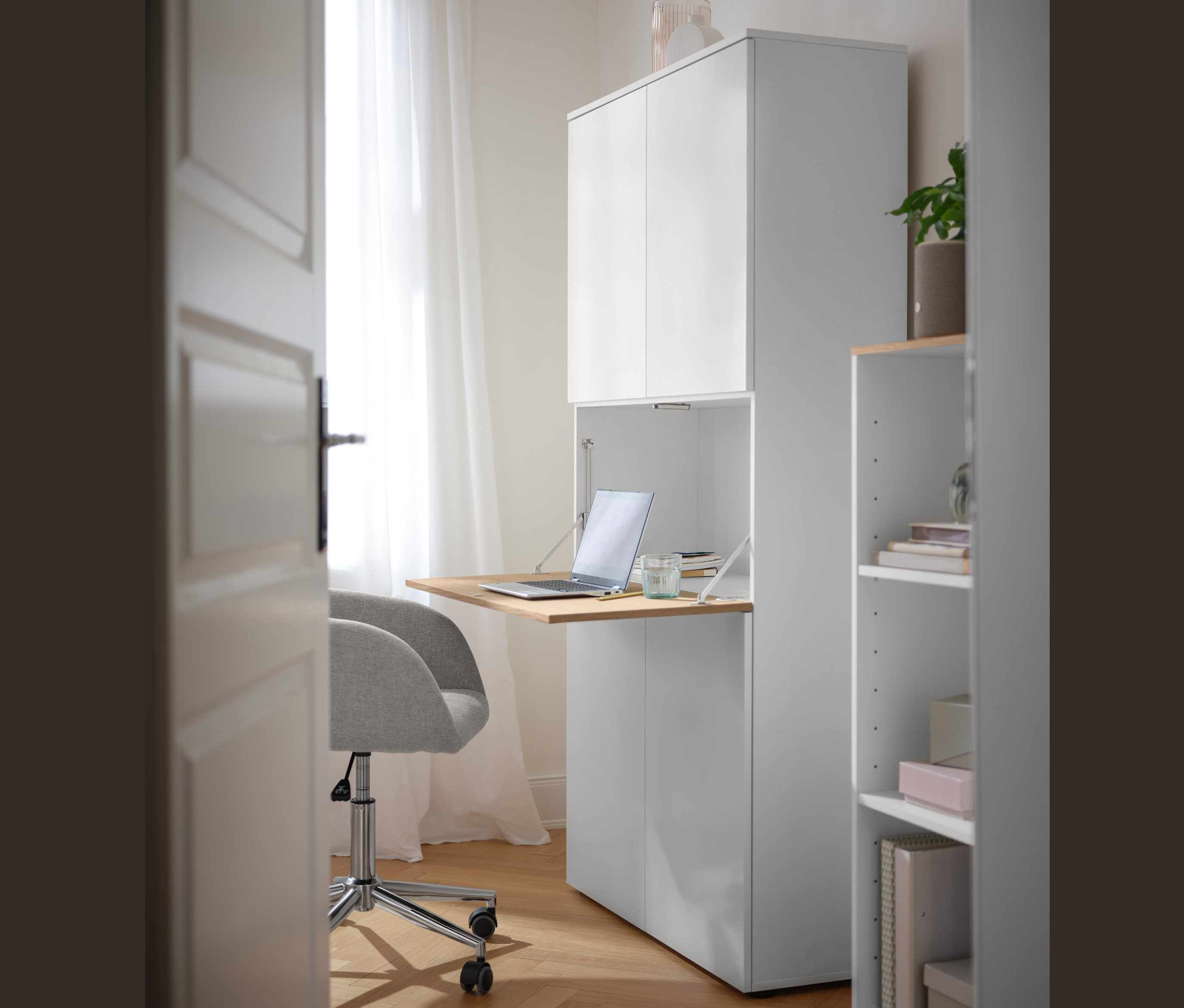 Beżowe krzesło do biurka stoi przy wysokiej białej szafce z blatem, na którym stoi laptop. Obok stoi szafka na kółkach.