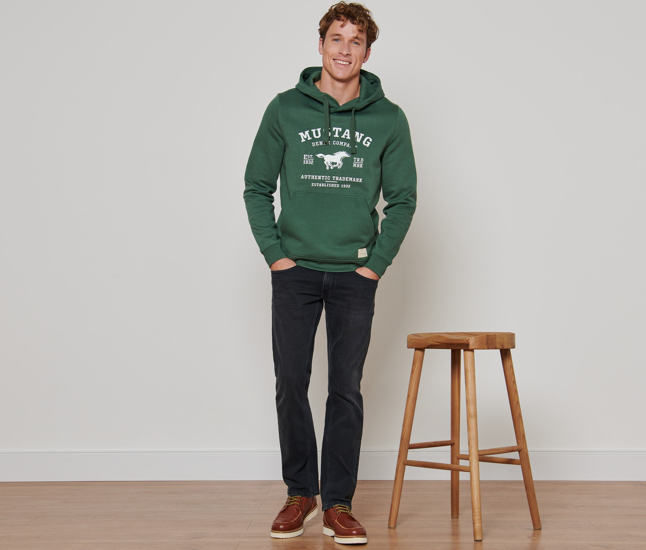 Mężczyzna stoi z rękami w kieszeniach szaro-czarnych spodni dżinsowych »Mustang«.