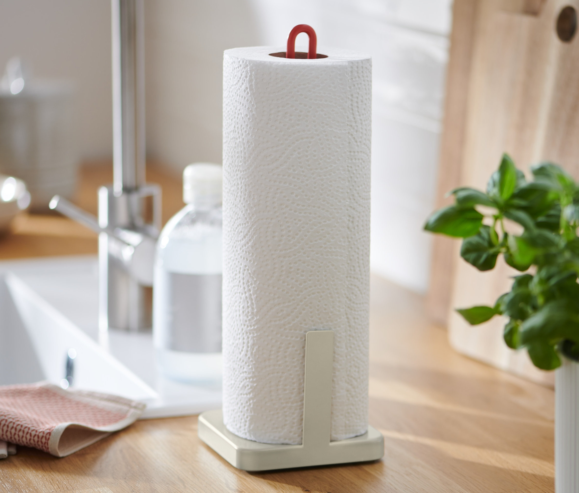 Na blacie kuchennym stoi uchwyt na ręczniki papierowe, zlew, dozownik mydła i doniczka z bazylią.