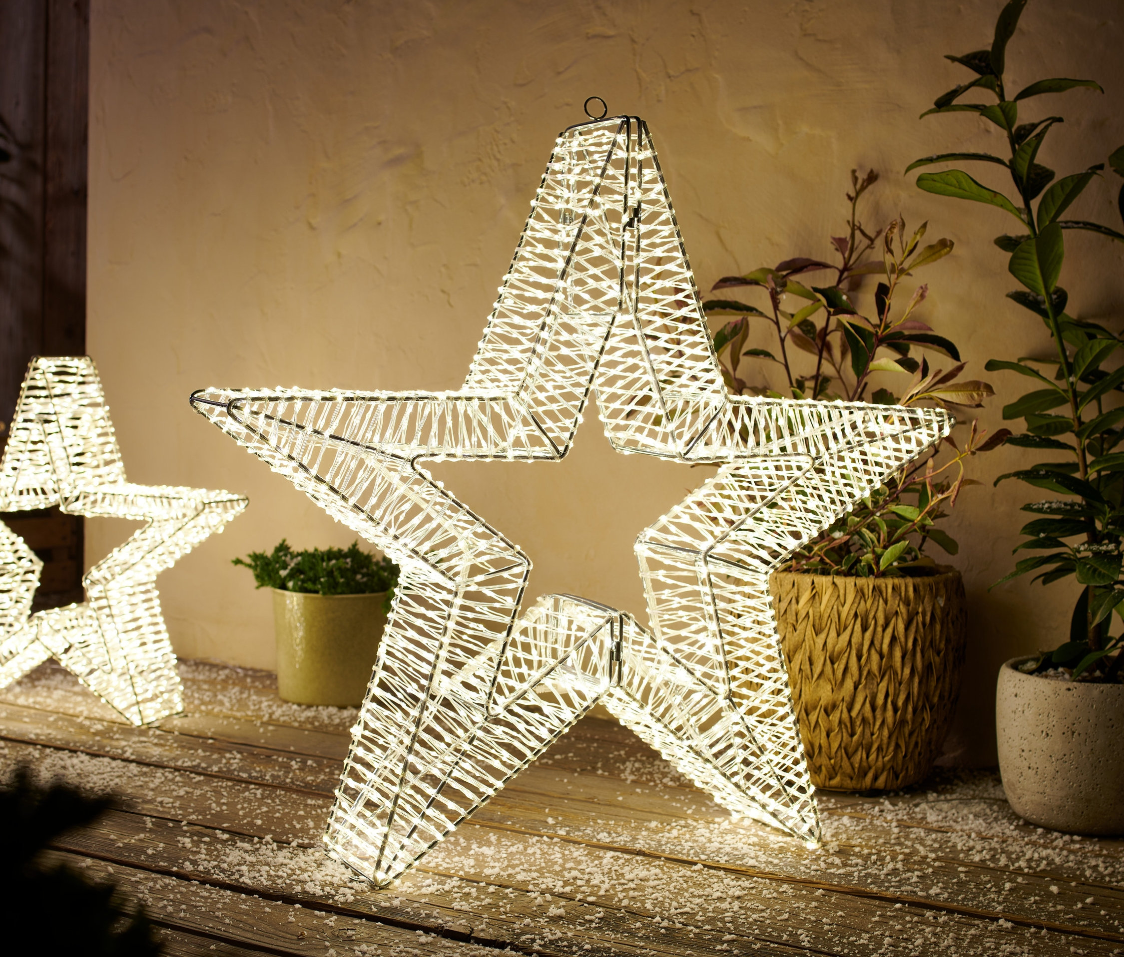 Dwie ogrodowe gwiazdy 3D LED, rozmiar L, stoją na drewnianej podłodze posypanej śniegiem. W tle rośliny w doniczkach.