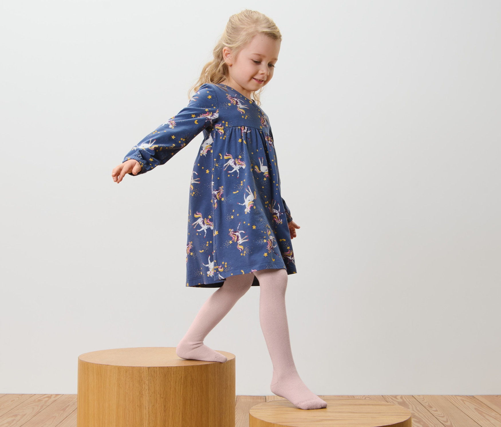Dziewczynka w niebieskiej sukience w jednorożce i różowych rajstopach bawełnianych, różowo-brązowe, 5 par, idzie po drewnianych stopniach.
