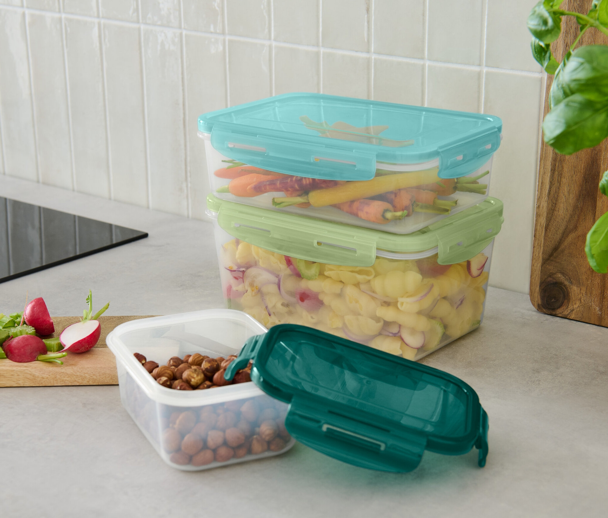 Na blacie kuchennym stoją trzy pojemniki na żywność: jeden z orzechami i zielonym wieczkiem, jeden z makaronem i zielonym wieczkiem oraz jeden z marchewkami i niebieskim wieczkiem.