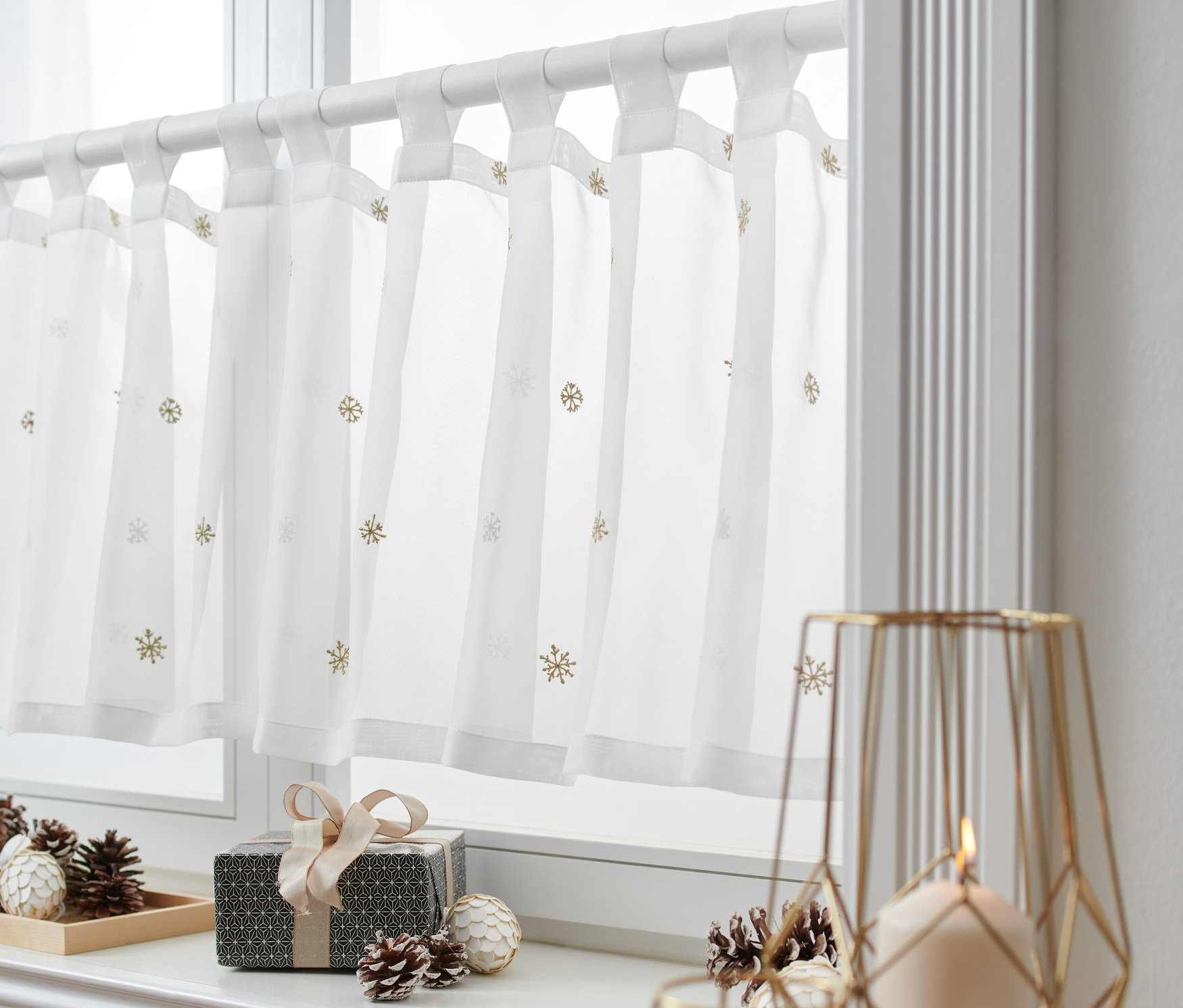 Biała zazdrostka z motywem płatków śniegu wisi w oknie. Na parapecie leżą pudełko z prezentem i szyszki.