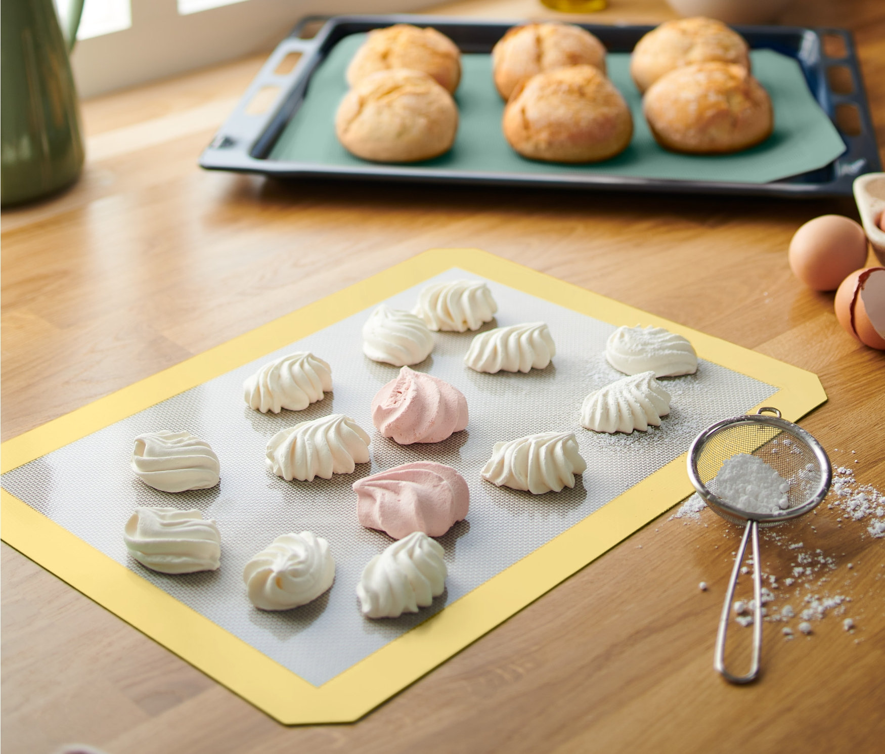 Na żółtej profesjonalnej macie do pieczenia leżą białe i różowe bezy. W tle blacha z bułkami.
