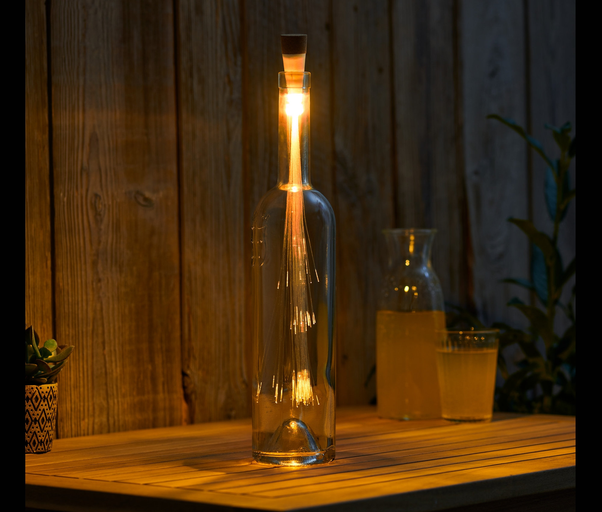 Na drewnianym stole stoi butelka z korkiem LED z akumulatorem, obok dzbanek i szklanka z napojem.