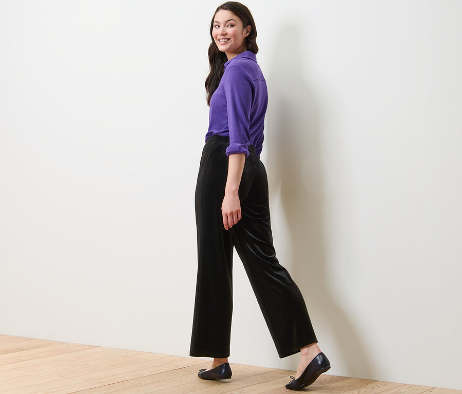 Kobieta idzie i spogląda przez ramię, ma na sobie fioletową koszulę i czarne aksamitne spodnie palazzo.
