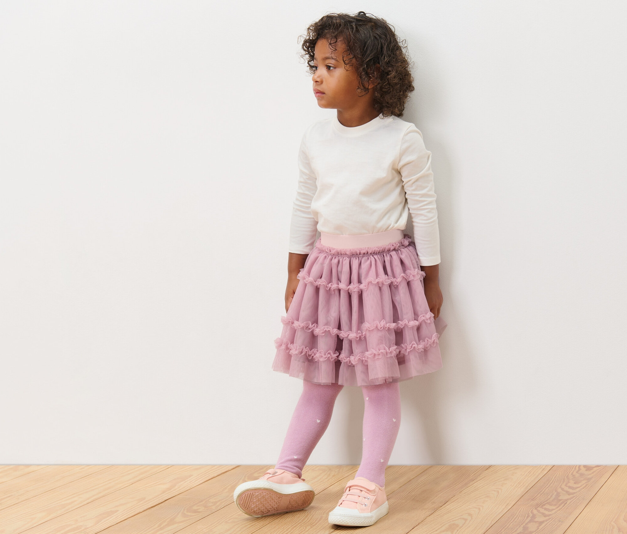 Dziewczynka w spódnicy tiulowej, bluzce z długim rękawem, legginsach i tenisówkach stoi.
