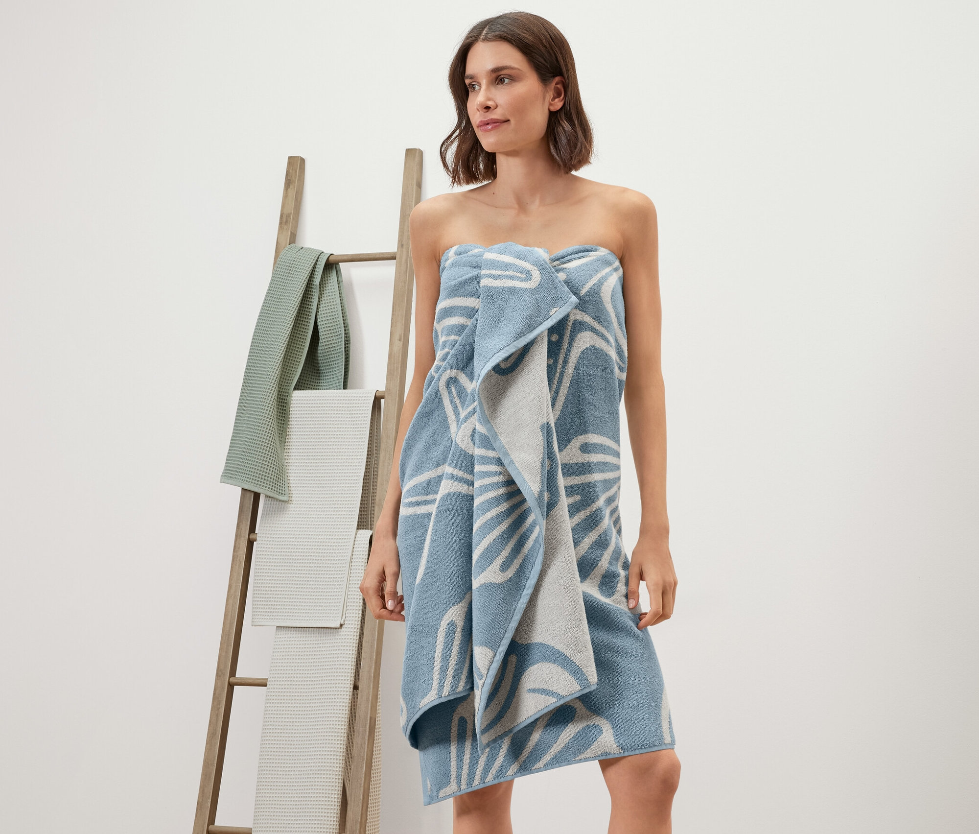 Kobieta owinięta ręcznikiem kąpielowym XL w muszle. W tle drewniana drabina z ręcznikami.