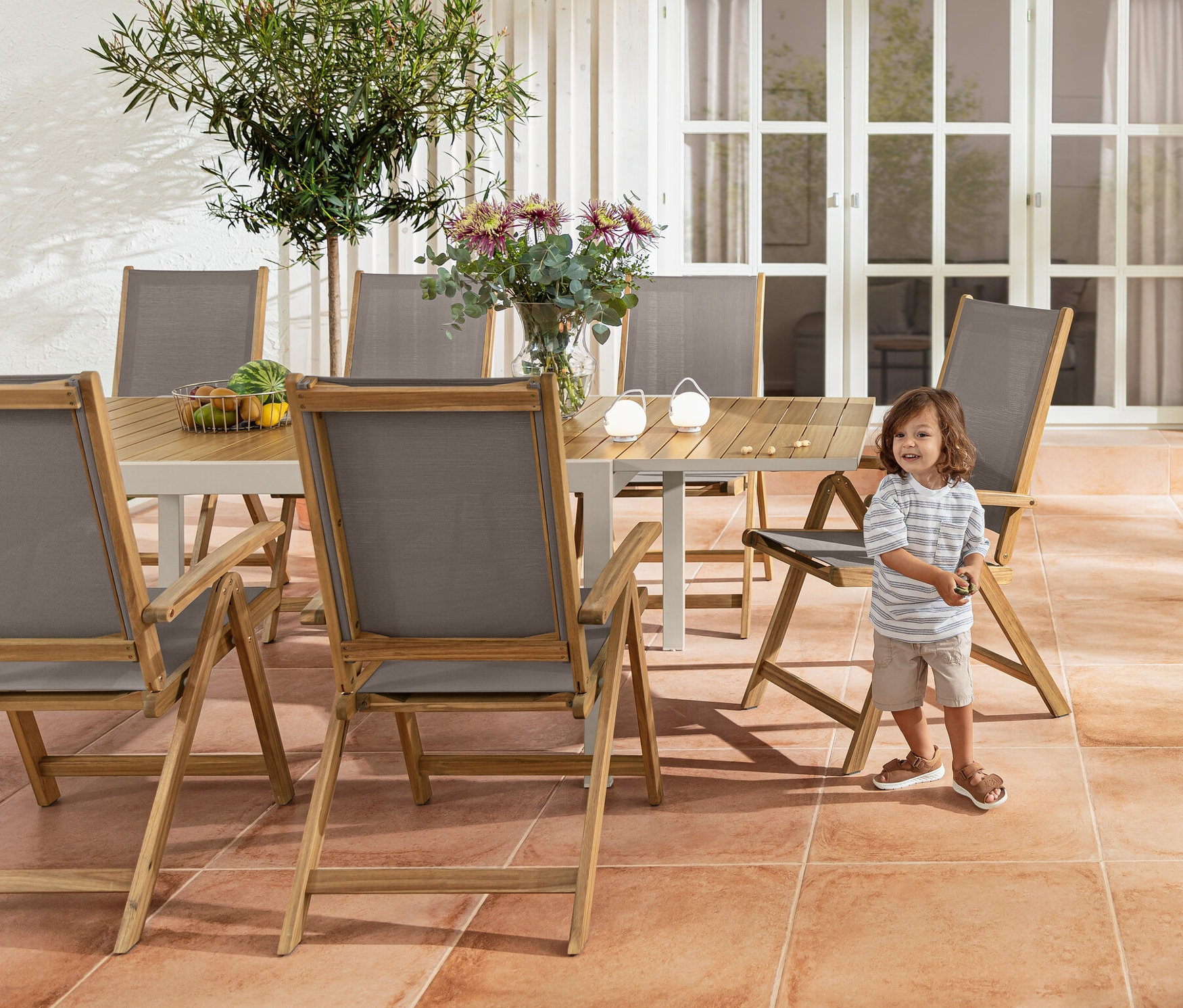 Chłopiec stoi obok rozkładanego stołu „Liska” z aluminium i krzesła ogrodowego „Lenja” z drewnianą ramą. W tle wózek barowy i zestaw akumulatorowych lamp stołowych.