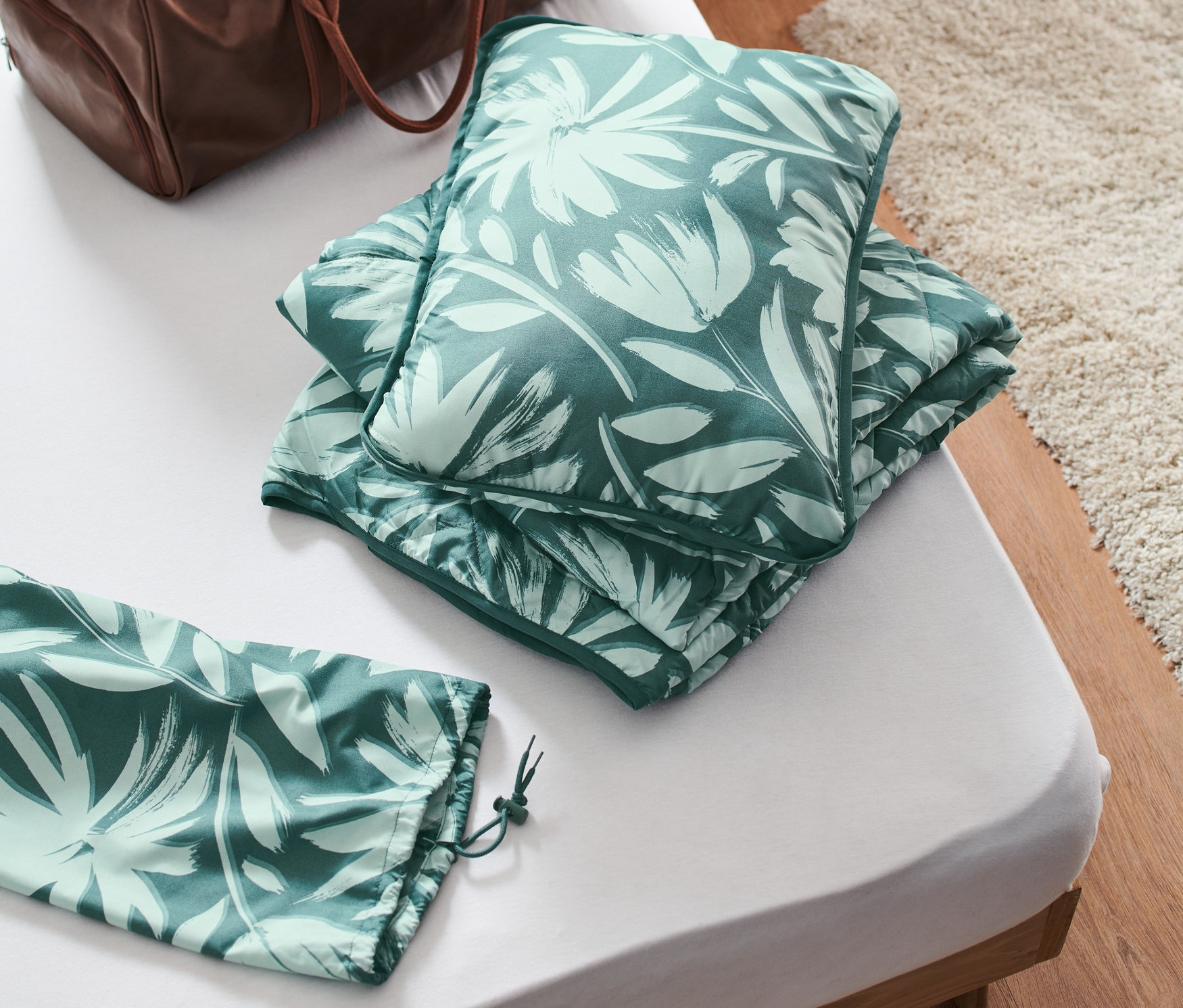 Na łóżku leży turystyczny komplet pościeli i brązowa torba podróżna.