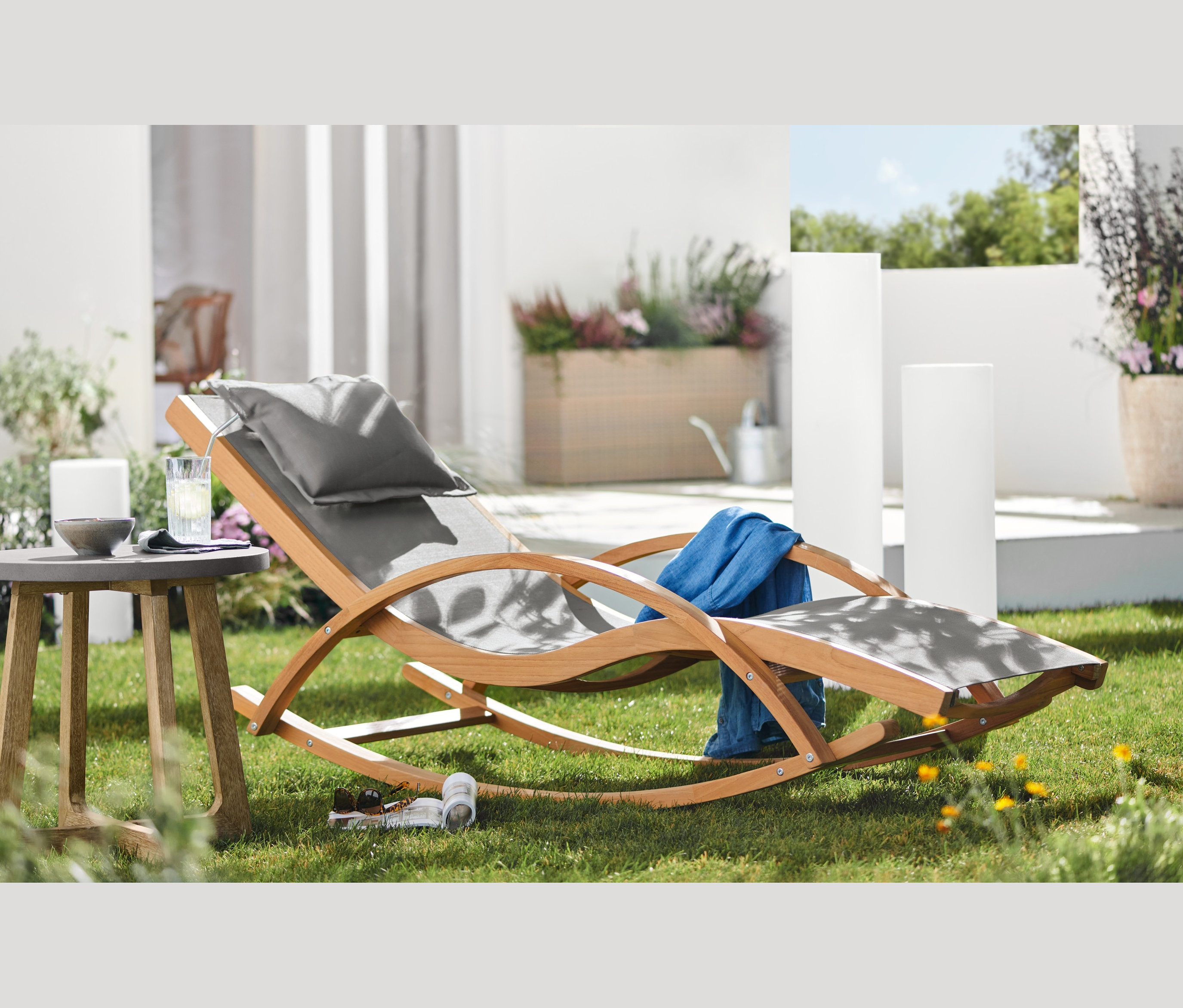 Leżak bujany z drewna tekowego z TEXTILENE® stoi na trawniku obok stolika z napojem.