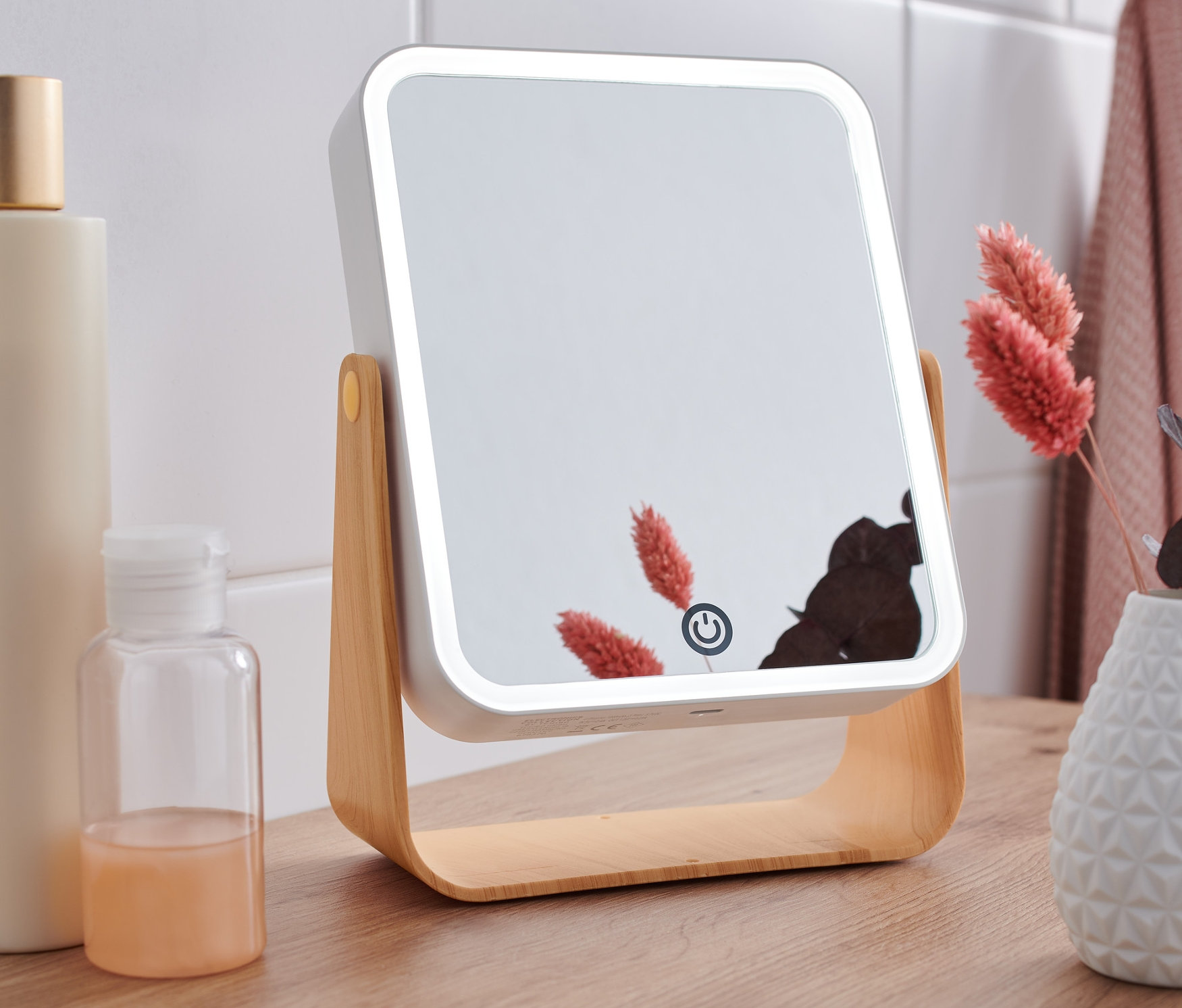 Na drewnianym stole stoi lusterko kosmetyczne z podświetleniem LED, obok butelek i wazonu z kwiatami.