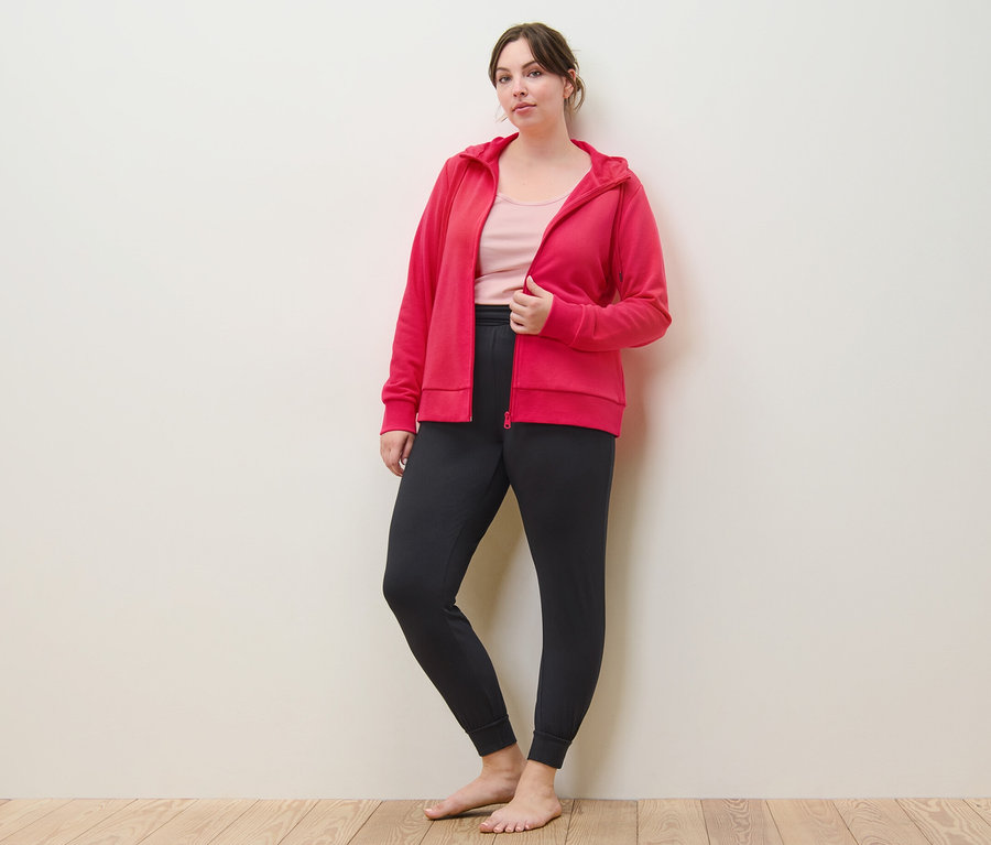 Kobieta pozuje w różowej rozpinanej bluzie dresowej, spodniach do jogi i topach (2 sztuki, bladoróżowy i różowy).