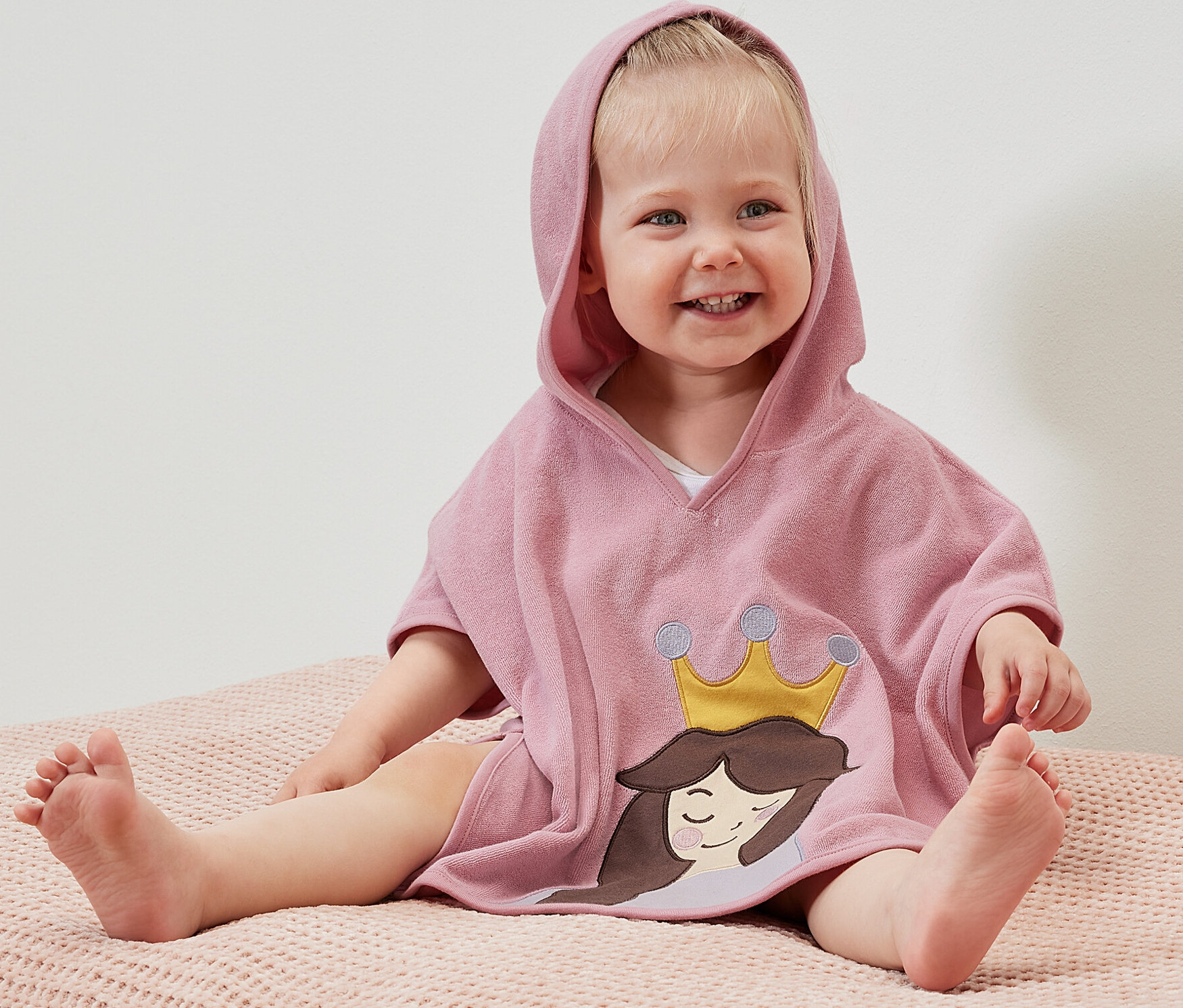 Uśmiechnięte dziecko siedzi na różowej powierzchni i ma na sobie ponczo z tkaniny frotté z kapturem z motywem księżniczki.