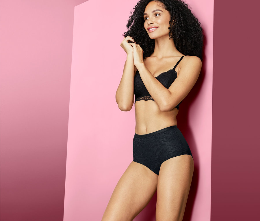 Kobieta o ciemnych, kręconych włosach pozuje w czarnych figach modelujących z wysokim stanem i koronkowym biustonoszu na różowym tle.