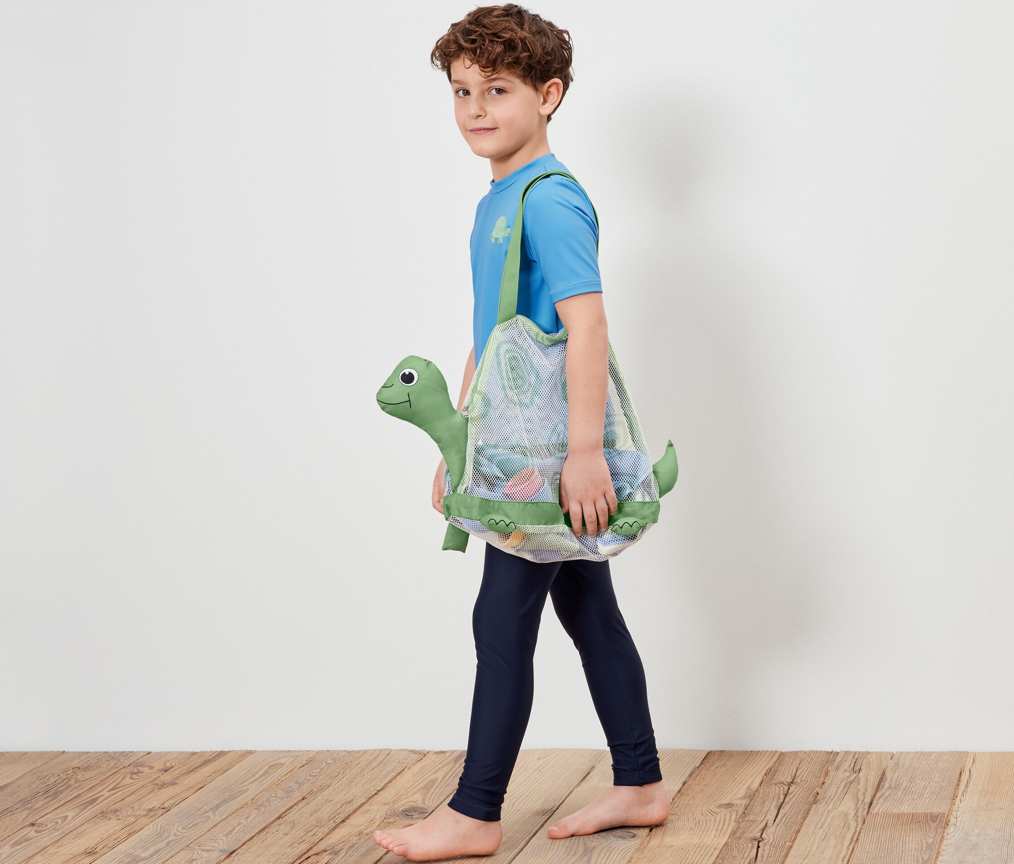 Chłopiec idzie i niesie siatkową torbę na zabawki plażowe z motywem żółwia.