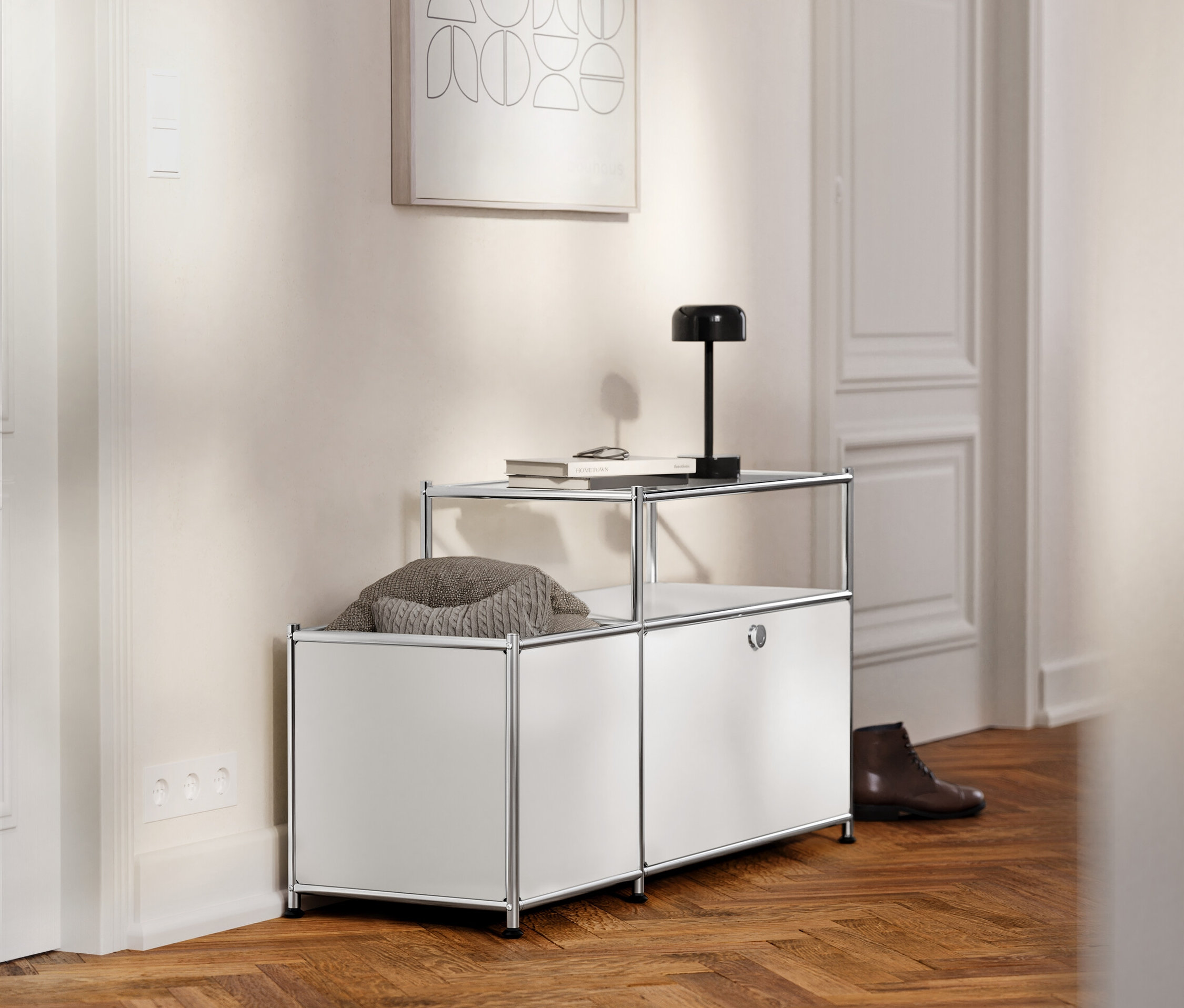 W korytarzu stoi biała metalowa szafka RTV »CN3« z klapą i szklanym blatem, na której leżą książka i lampa.